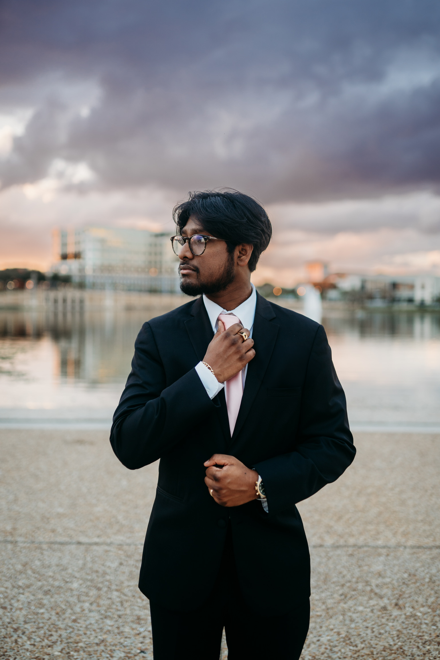 A man in a black suit adjusting his light pink tie outdoors near water with city buildings and cloudy sky in the background.