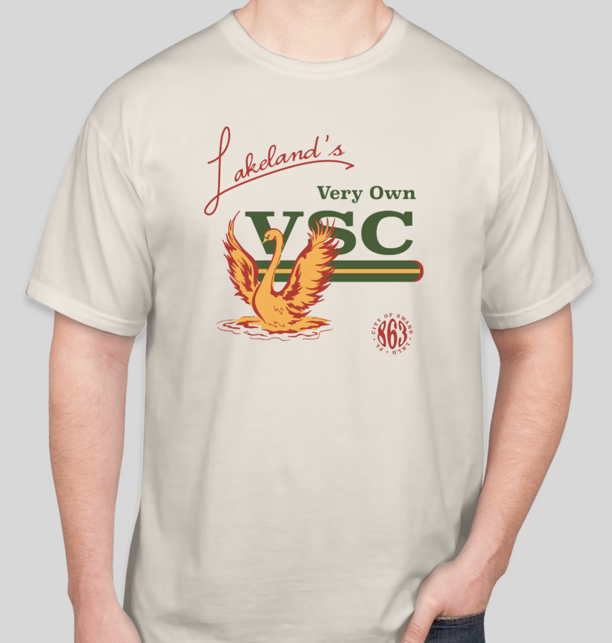 A person wearing a beige T-shirt with a graphic design featuring a flamingo, the text 'Lakedland's Very Own WSC,' and a red circle with the number '863' and the words 'City of Swans.'
