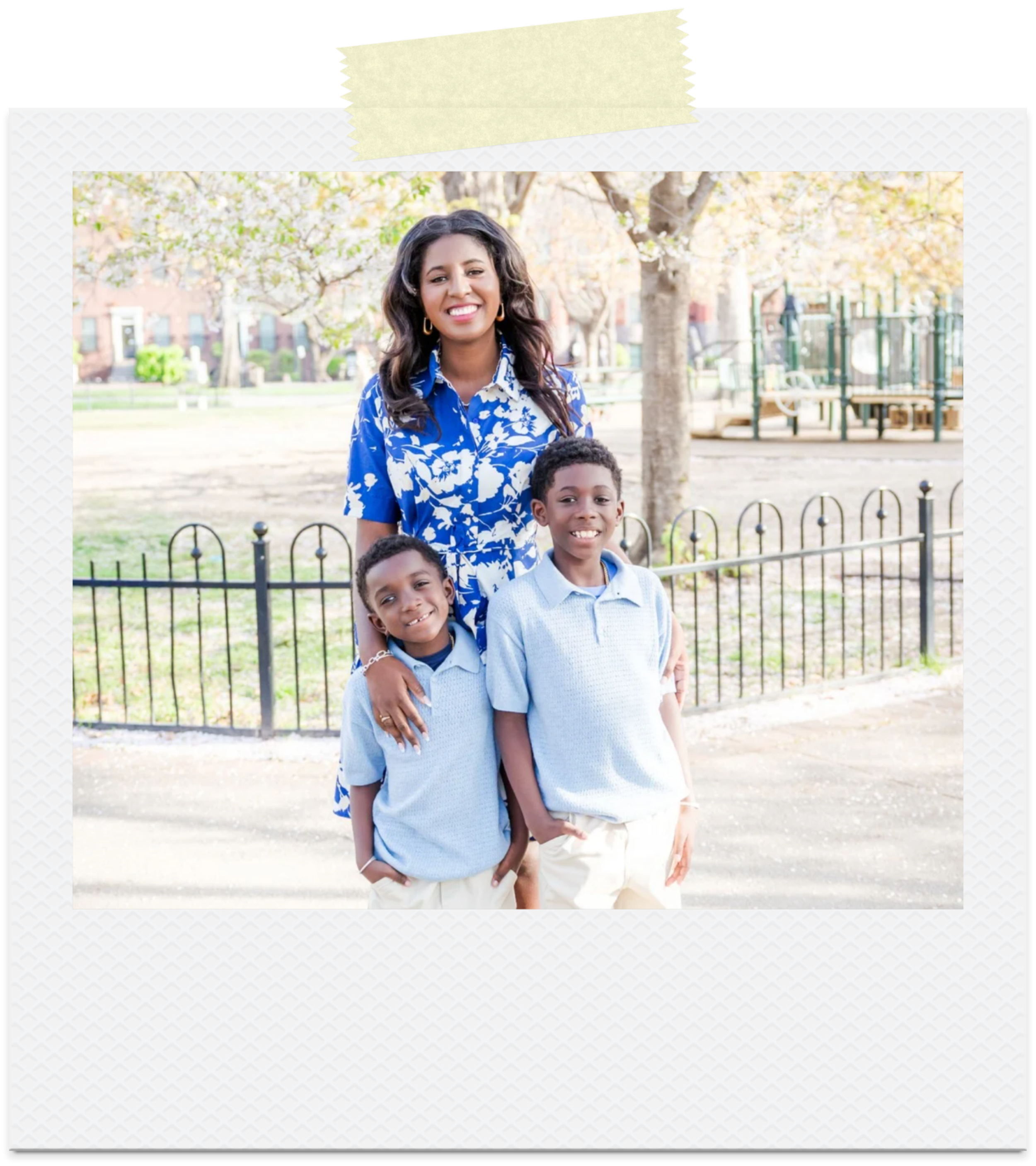 Smiling woman with curly dark hair in a floral blue blouse and three boys in light blue shirts standing on a sidewalk in a park with trees and playground equipment in the background.