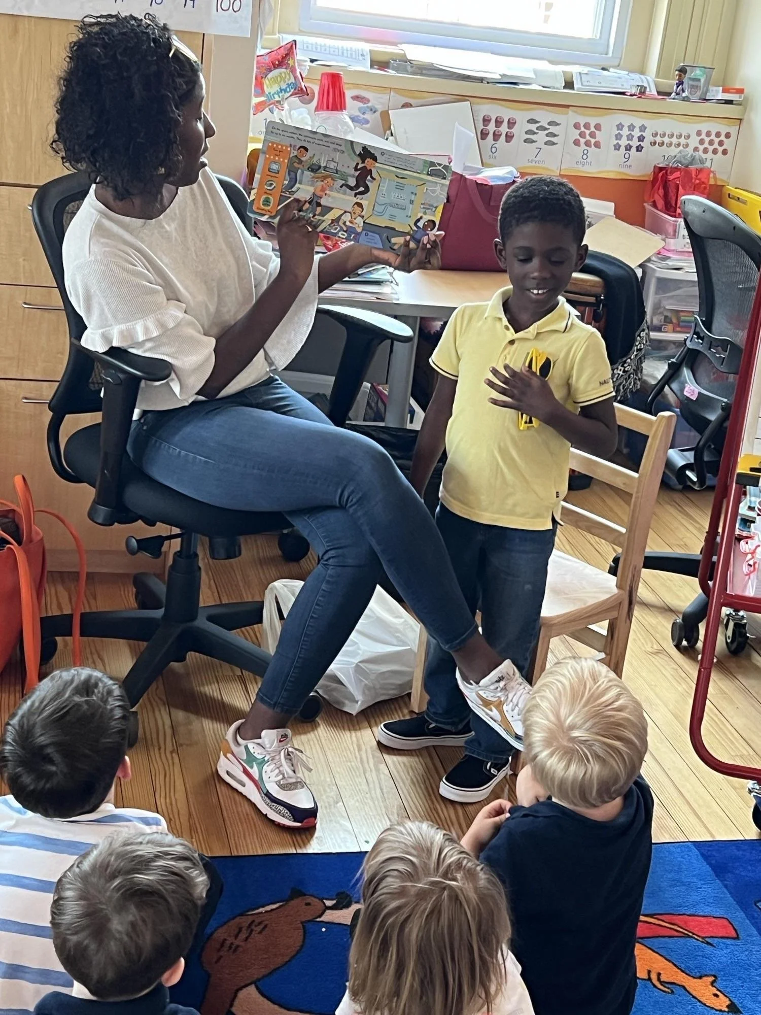 A woman reading a children's book to a group of children sitting on the floor in a classroom.