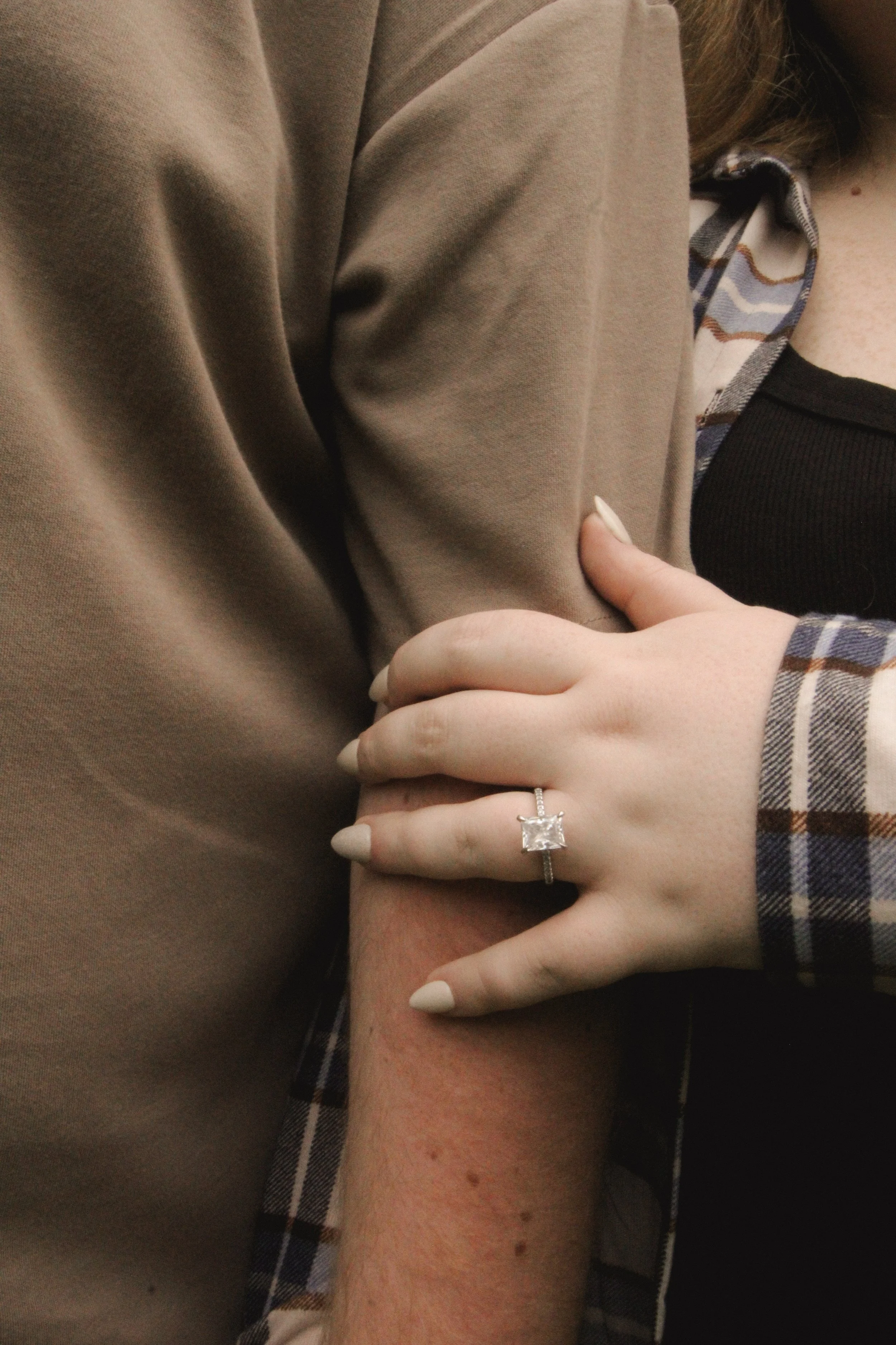 Close-up of a woman’s hand with a large square-cut diamond engagement ring on her finger, resting on a man's arm. The man is wearing a brown jacket and the woman is wearing a plaid shirt.