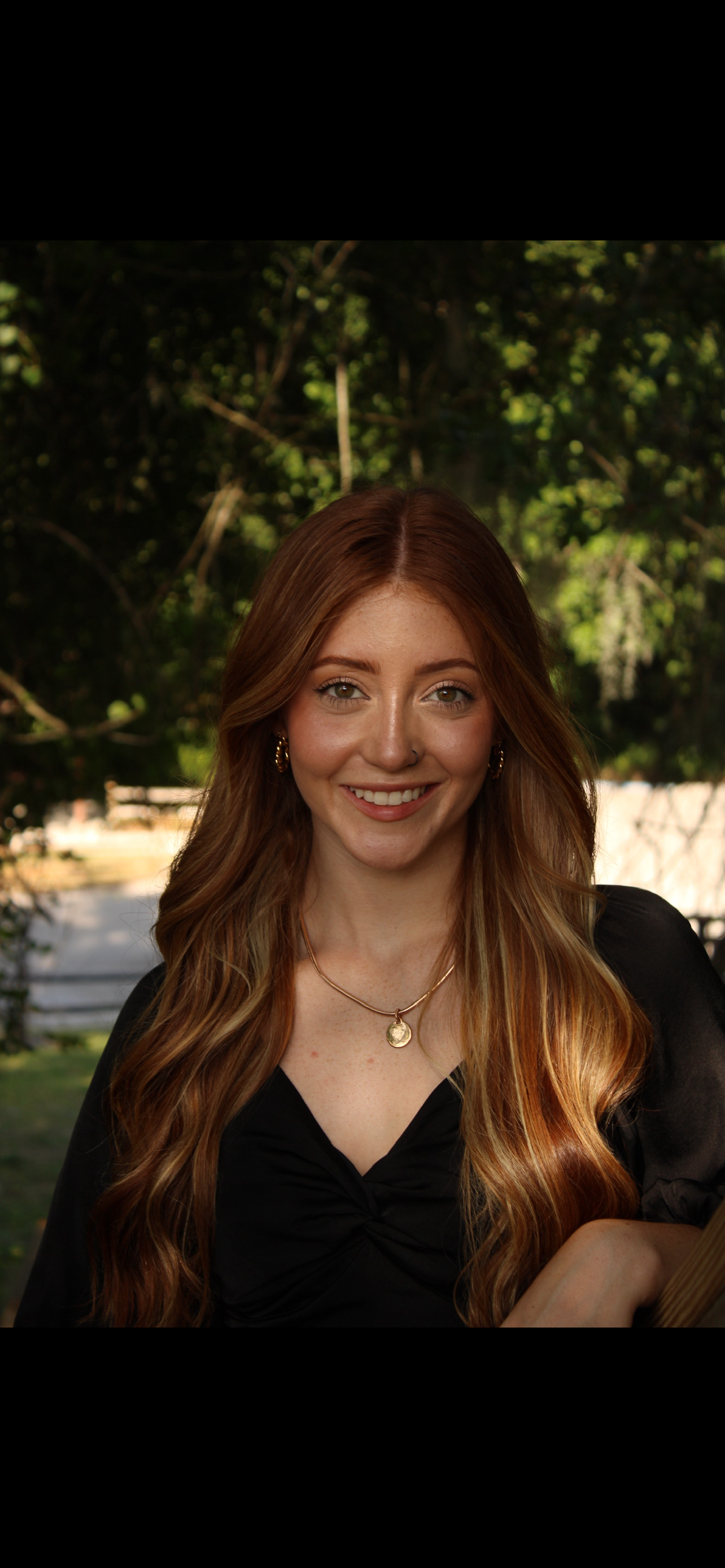 Portrait of a young woman with long red hair, wearing earrings, a necklace with a round pendant, and a black top, smiling outdoors with trees in the background.