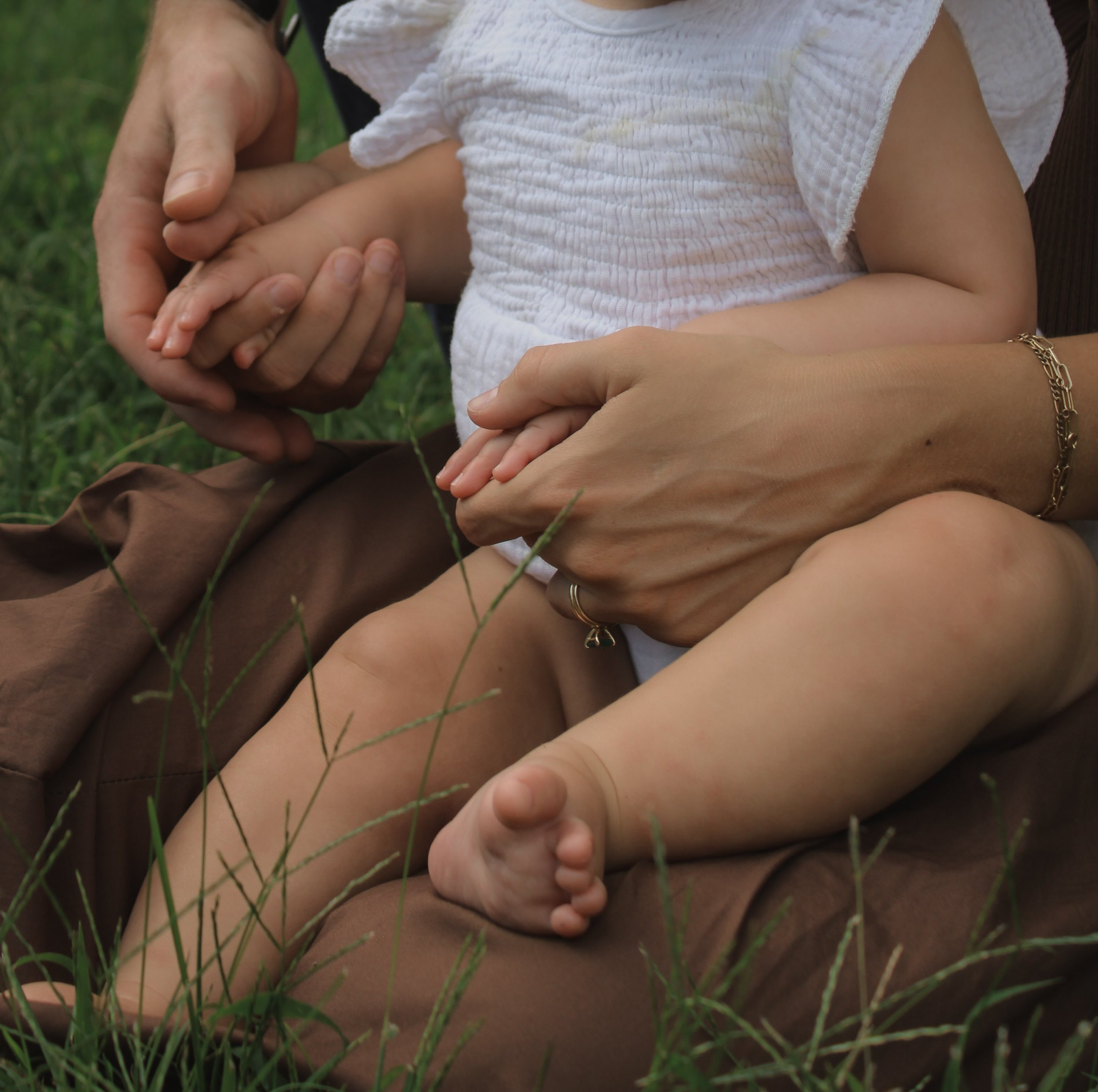 A close-up of a child's arms being held and embraced by multiple adults outdoors, with green grass visible at the bottom of the image.