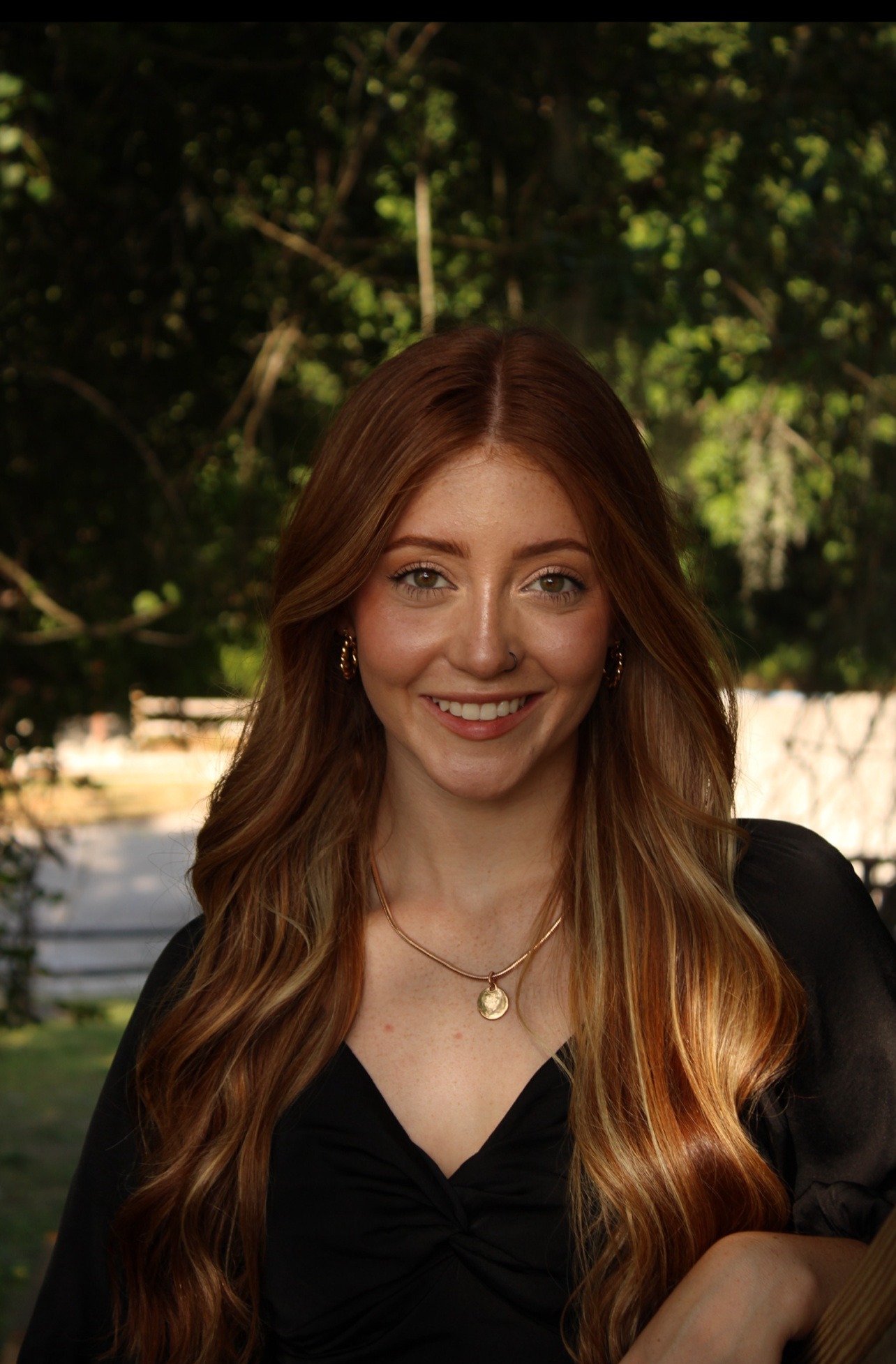 A young woman with long, wavy red hair, wearing gold hoop earrings, a gold necklace with a pendant, and a black top, smiling outdoors with lush green trees in the background.