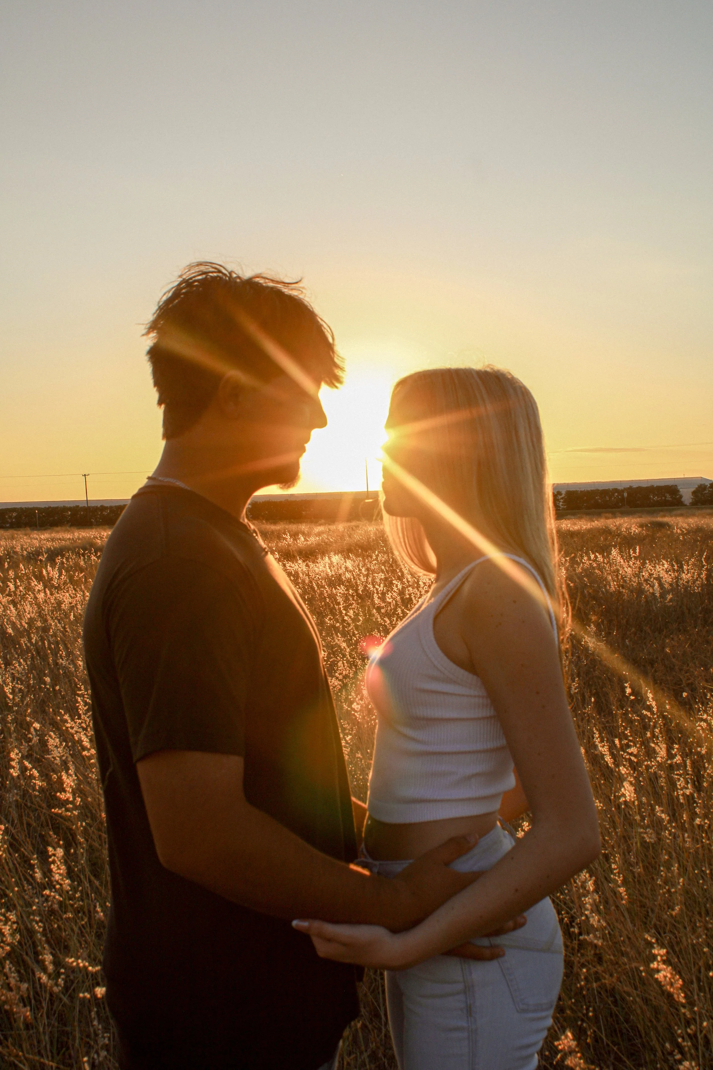 A man and woman stand close in a field during sunset, facing each other with sunlight shining behind them, creating a warm, romantic atmosphere.