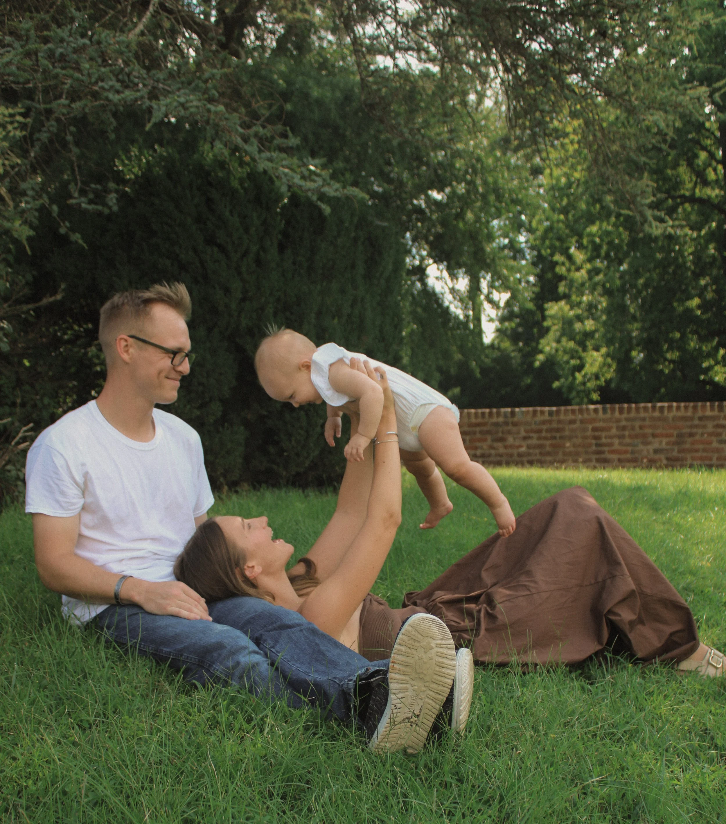 A family of three enjoying outdoor time in a park. The father is sitting on the grass with a woman lying behind him, holding a child up in the air. The setting includes lush green grass, trees, and a brick wall in the background.