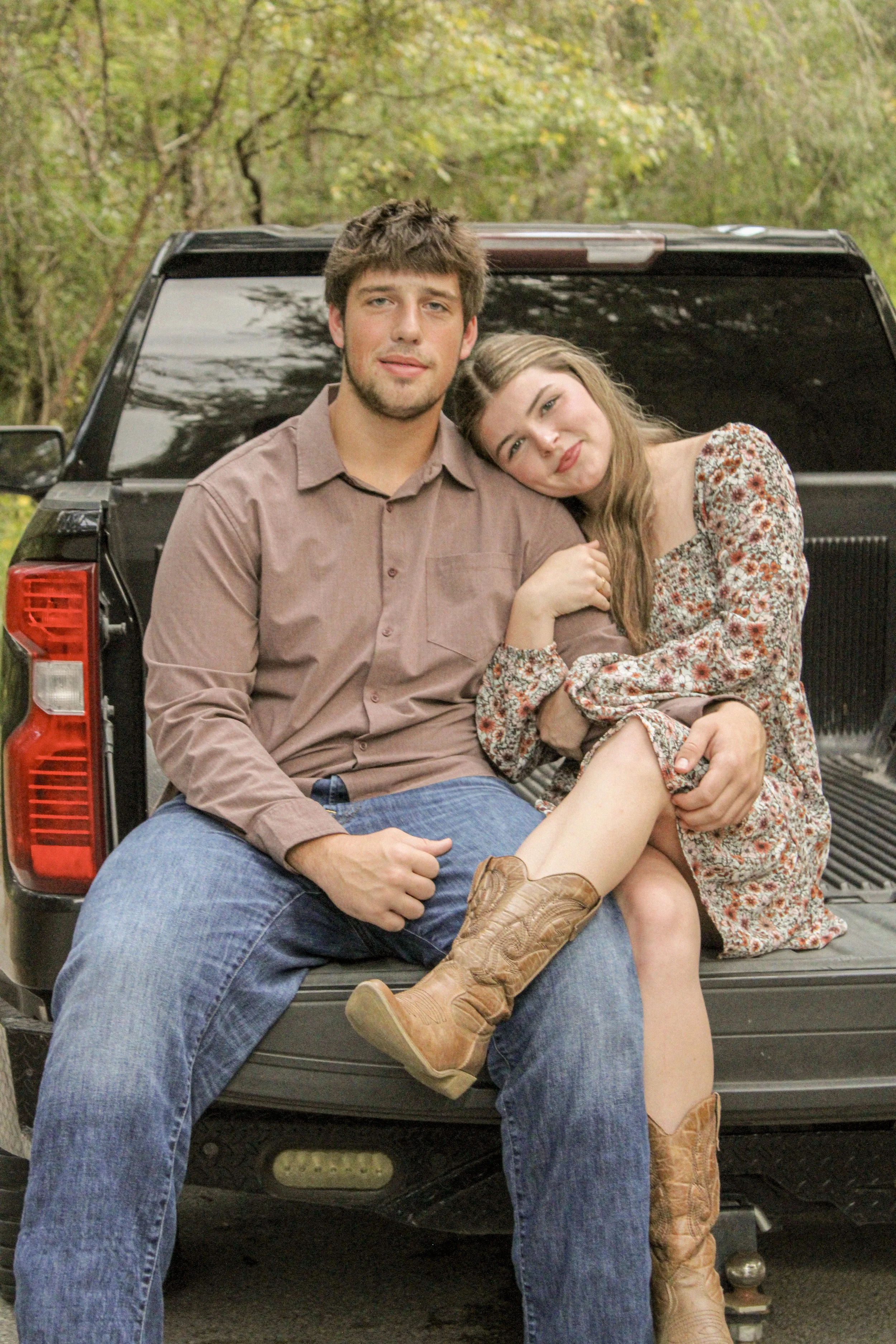 A young man and woman sitting on the tailgate of a black truck in a wooded outdoor area. The woman is resting her head on the man's shoulder, and they are both looking at the camera. The woman is wearing a floral dress and cowboy boots, and the man is wearing a button-up shirt and jeans.