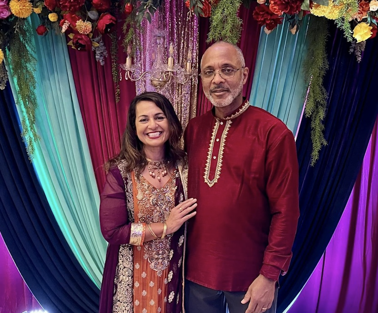 A smiling woman and a man dressed in traditional Indian attire at a colorful decorated event backdrop with flowers and drapes.