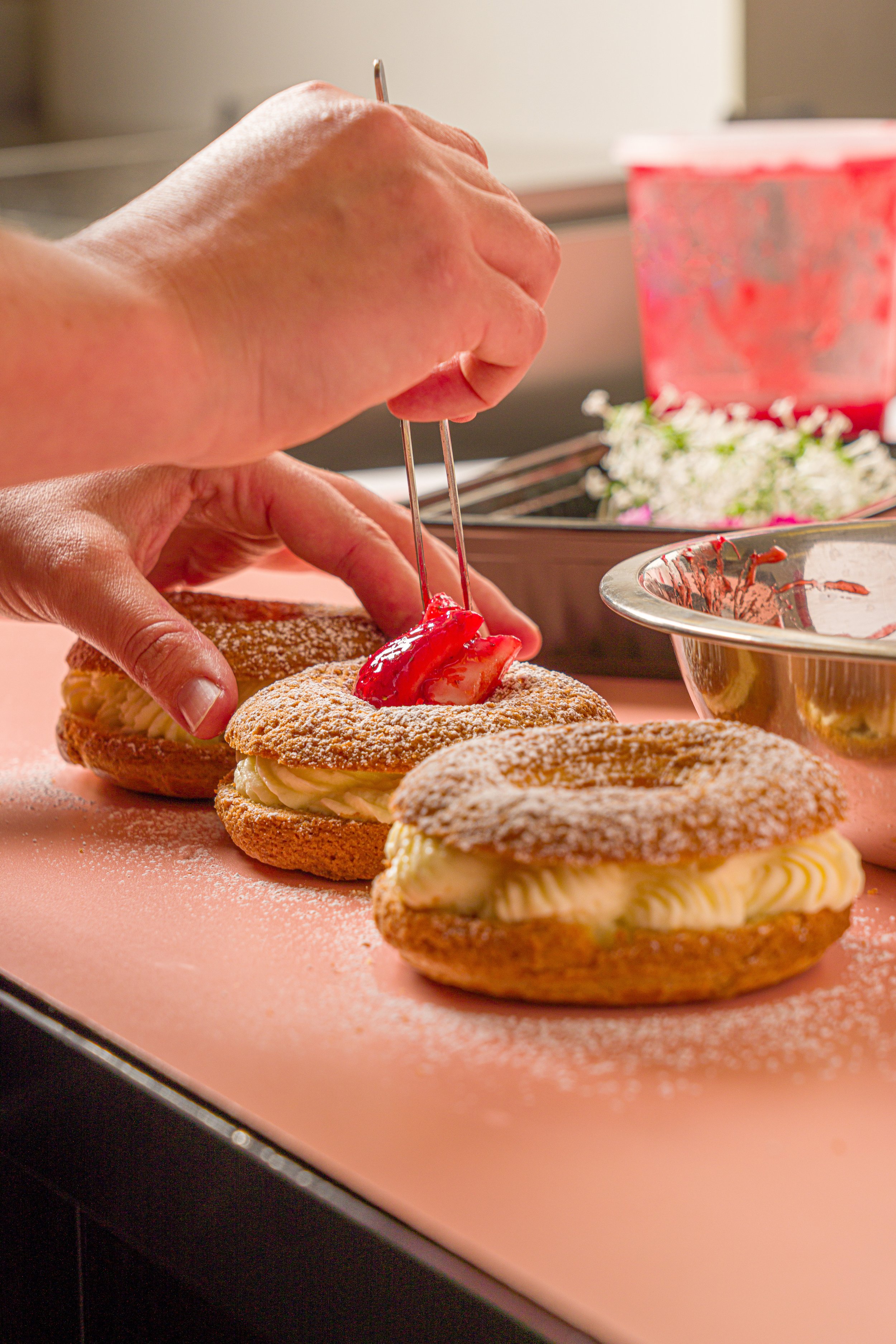 Close-up of a person's hand using tweezers to place a red cherry on top of a baked pastry with powdered sugar and cream filling, arranged on a pink surface with some flowers and a pink container in the background.