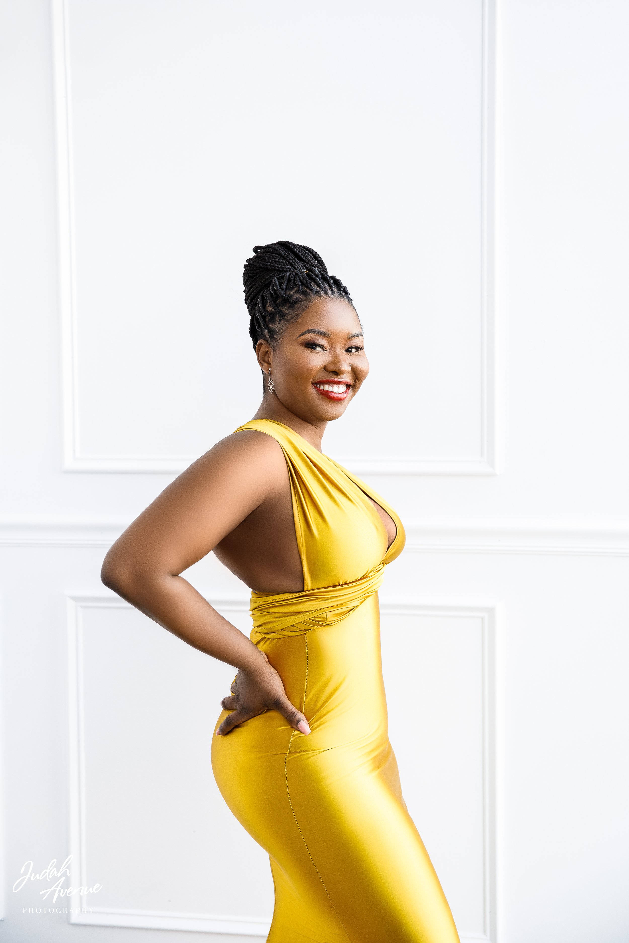 A woman in a shiny gold dress with a deep V neckline, styled in an elegant updo with twists, smiling confidently against a white wall with decorative molding.