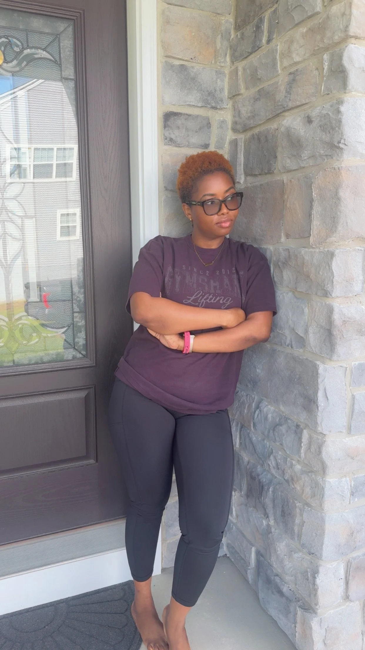A woman with short, curly blonde hair wearing glasses with a dark frame, a purple T-shirt with text, black pants, and a pink wristband, standing with arms crossed on a porch next to a stone wall and a dark door with decorative glass.