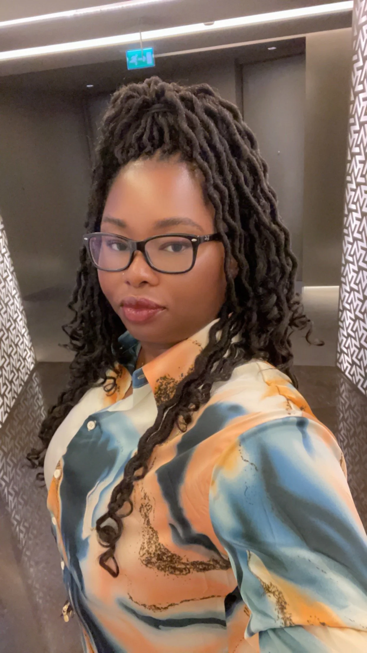 A woman with dark, curly, styled hair and glasses taking a selfie in an elevator with patterned walls and an exit sign above.