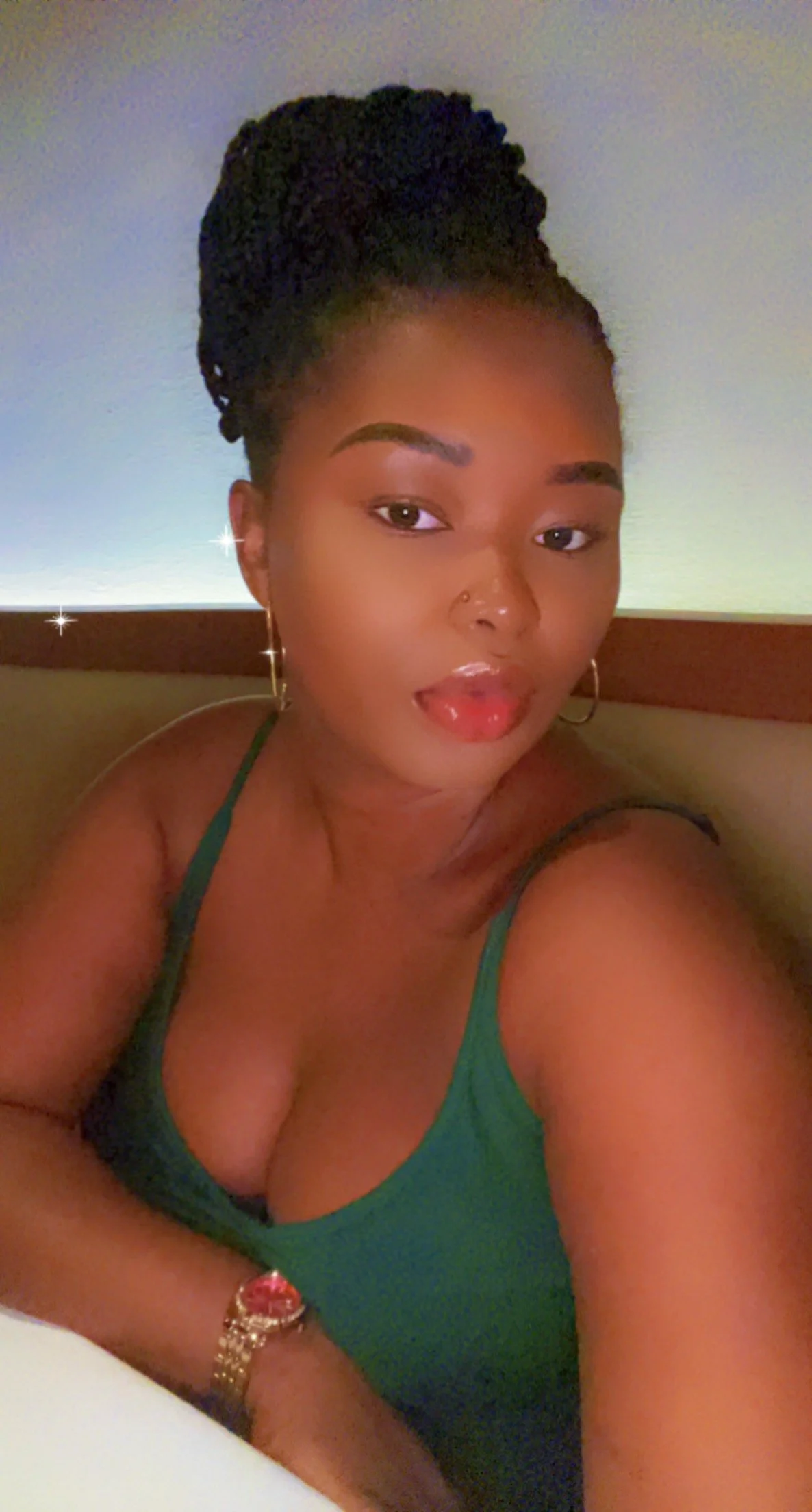 A woman with dark hair styled in dreadlocks tied up, wearing a green tank top, earrings, a wristwatch, and a nose ring, taking a selfie indoors with a plain wall background.