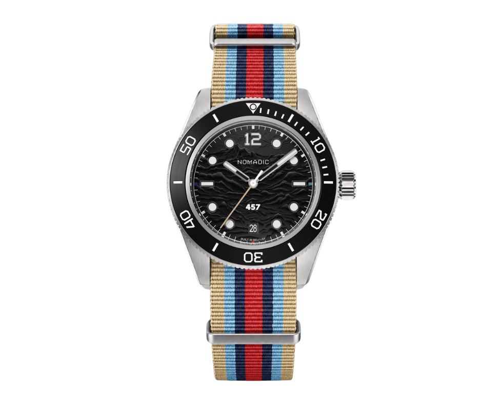 A wristwatch with a black dial featuring the brand name 'Nomadic', a date window, and a striped fabric strap in red, blue, tan, and black.