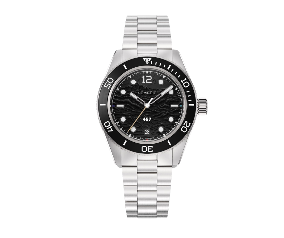 A silver wristwatch with a black bezel, black textured dial, white hour markers, and a date window at 6 o'clock. The watch has three hands and the word 'NOMADIC' on the dial.