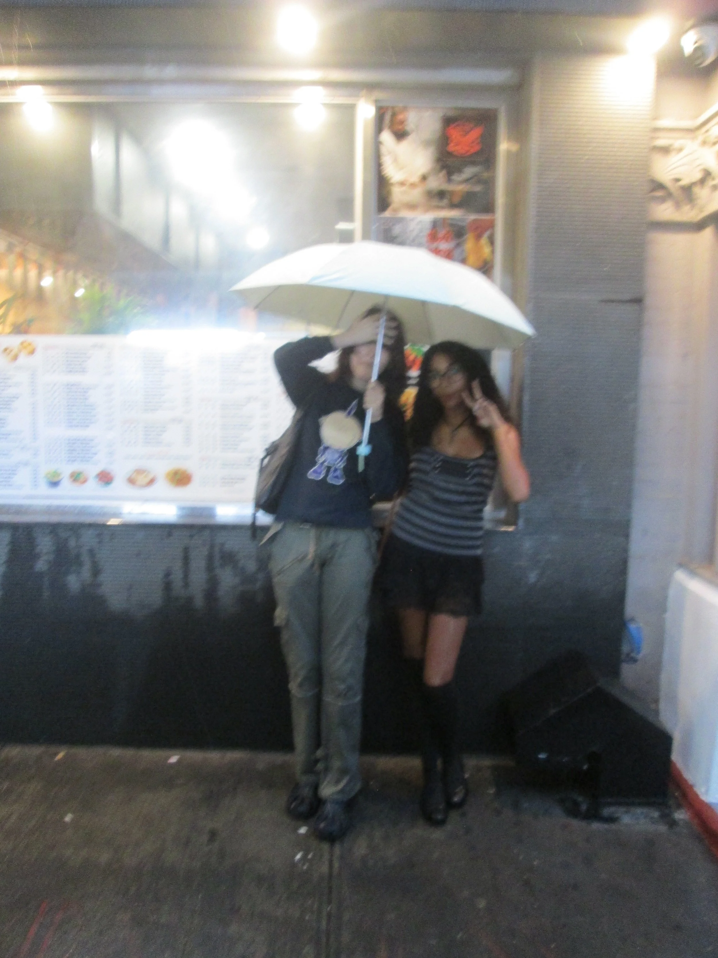 Two women standing close together outside, one holding a white umbrella over both of them. The woman on the left is wearing a dark jacket and pants, and the woman on the right is wearing a striped shirt, black shorts, and glasses. They are posing for