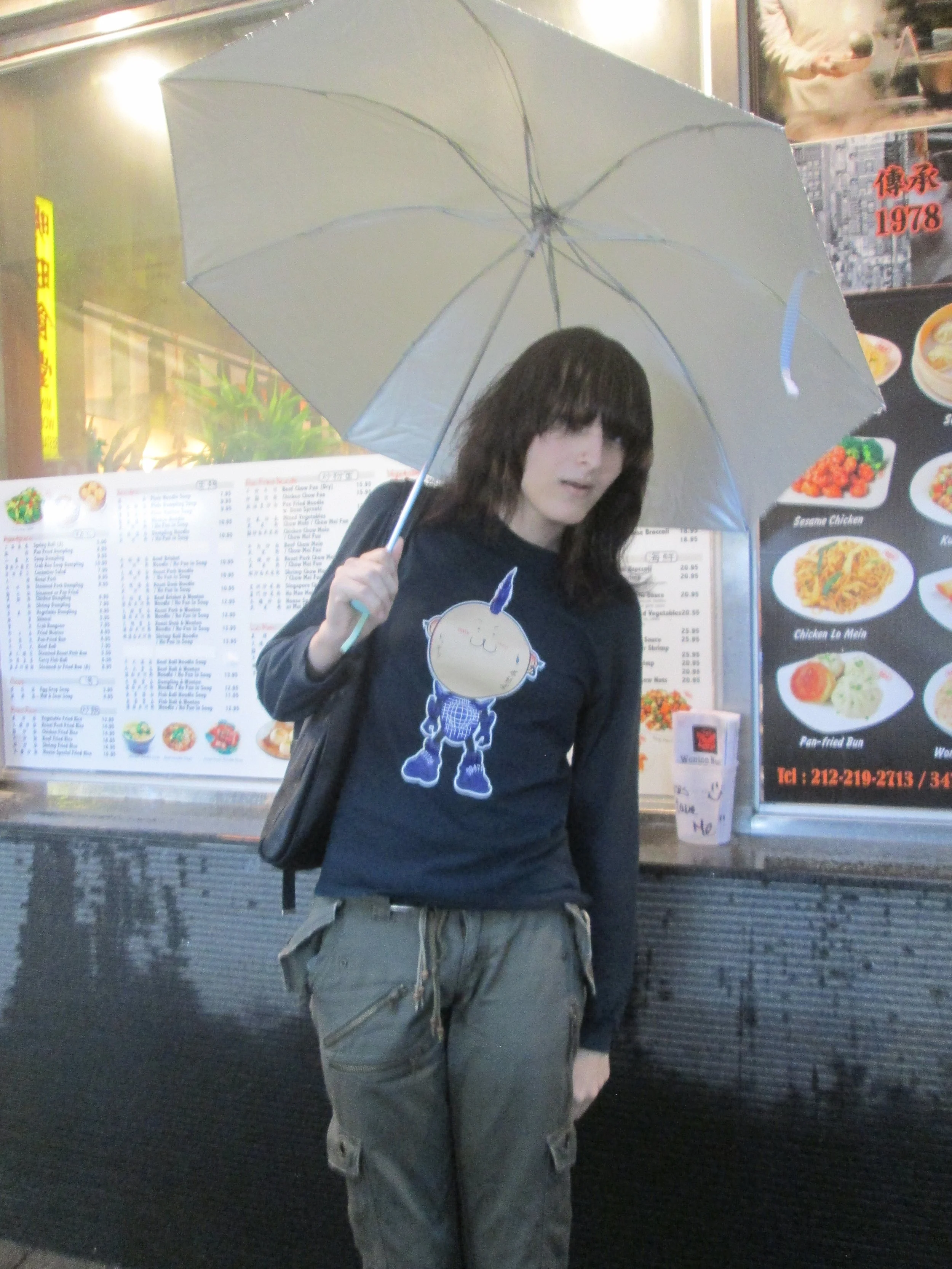 A woman holding a gray umbrella, standing in front of a restaurant menu board. She is wearing a navy blue long-sleeved shirt with a cartoon character on it and olive green cargo pants.