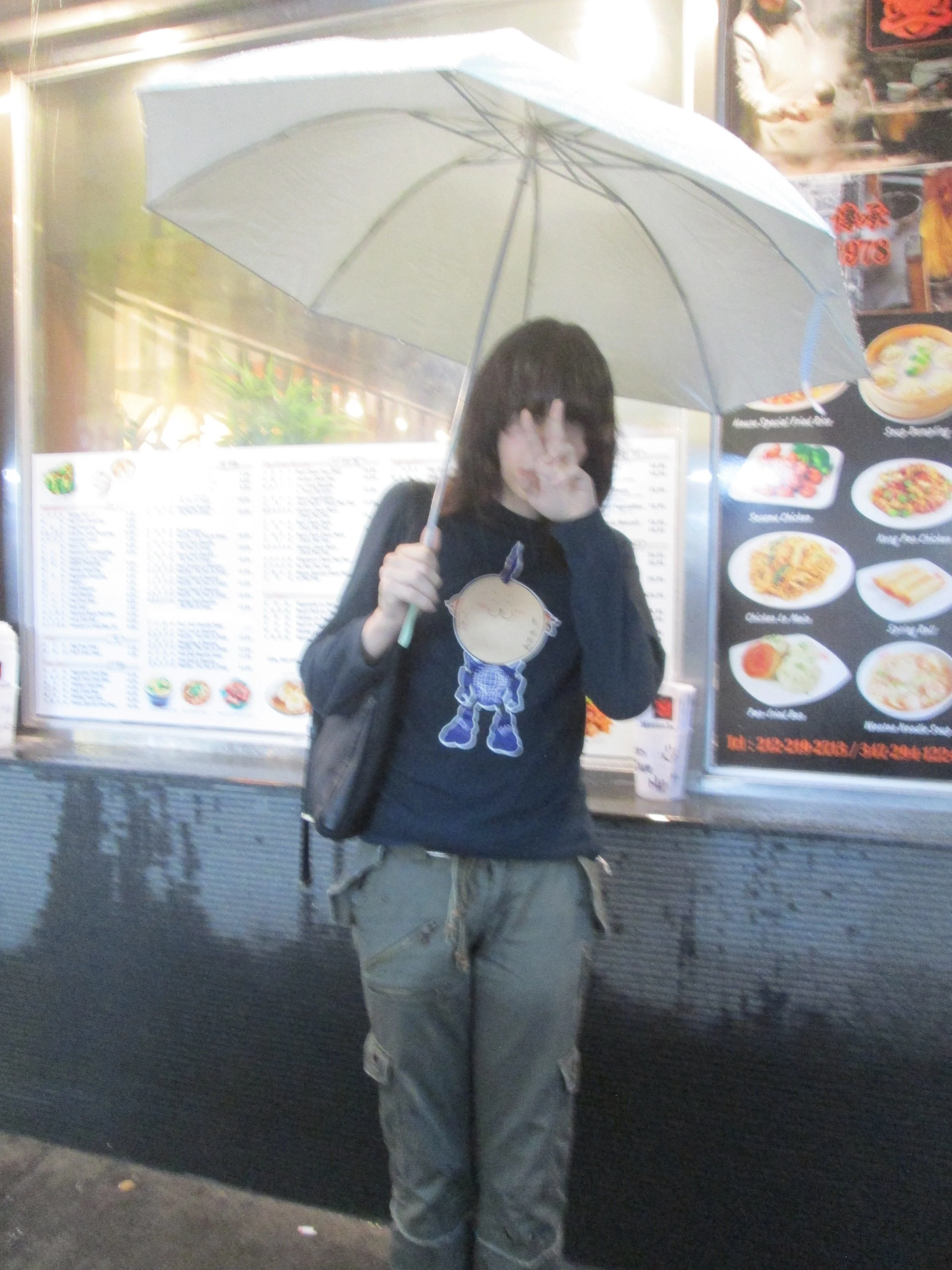 Person with dark hair holding a light-colored umbrella, standing in front of a menu board at a restaurant, wearing a black hoodie with a cartoon character and tan pants.