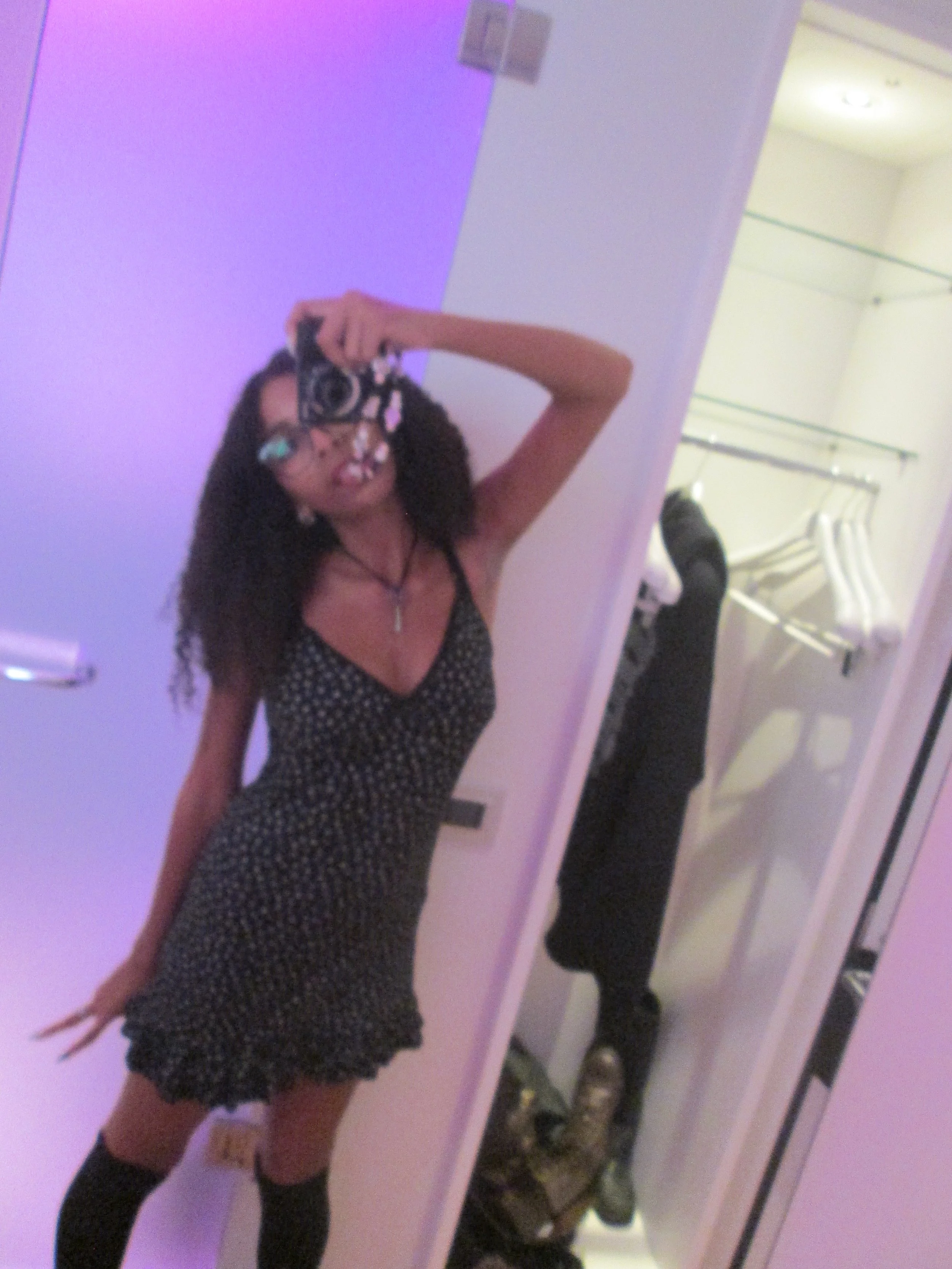 A woman taking a mirror selfie in a room with purple lighting, wearing a black polka dot dress and thigh-high socks.