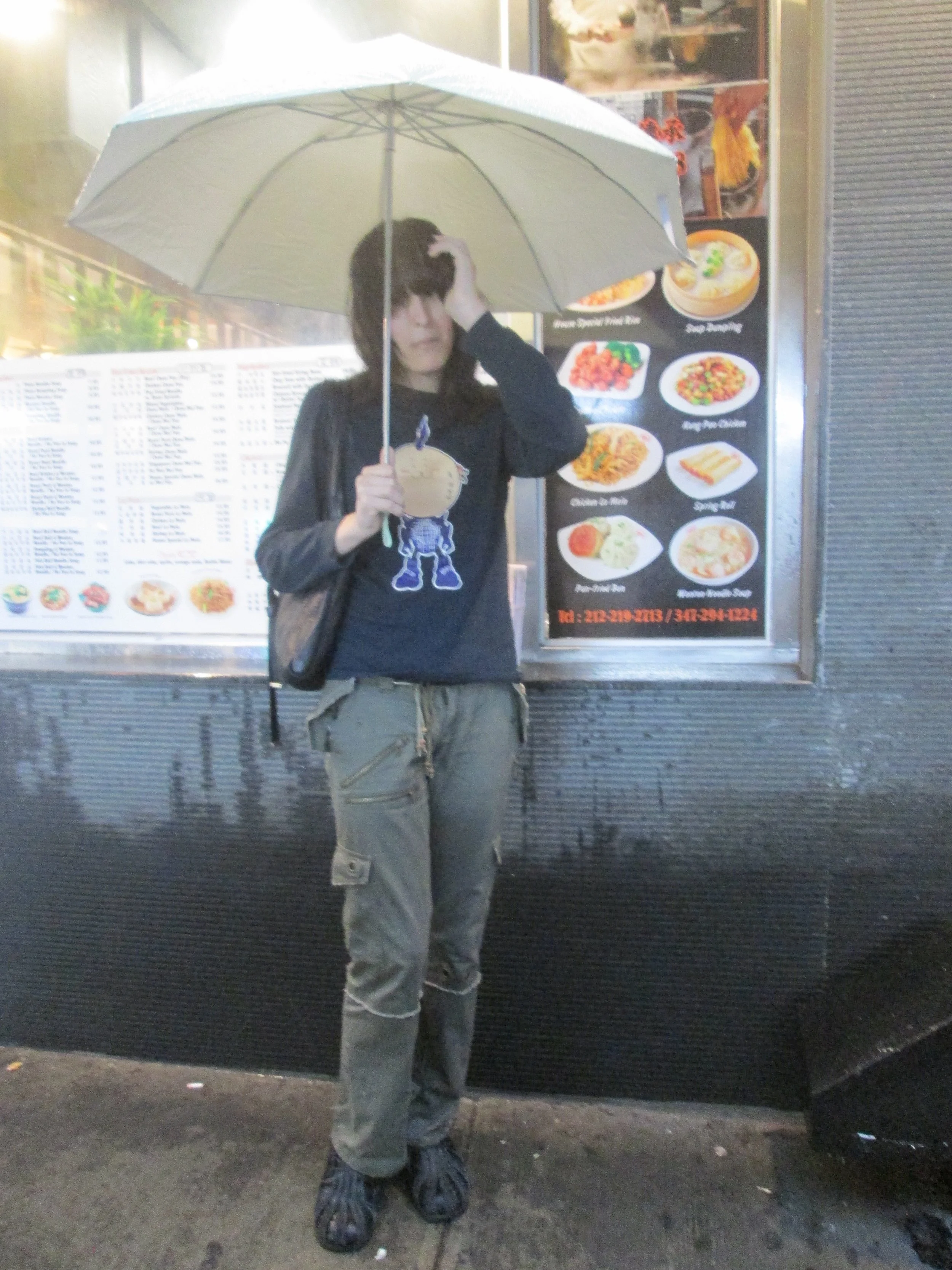 A person standing outside holding an open light-colored umbrella. The person is wearing a black jacket, khaki cargo pants, and sandals. Behind them is a menu display with food pictures and a black wall or panel.