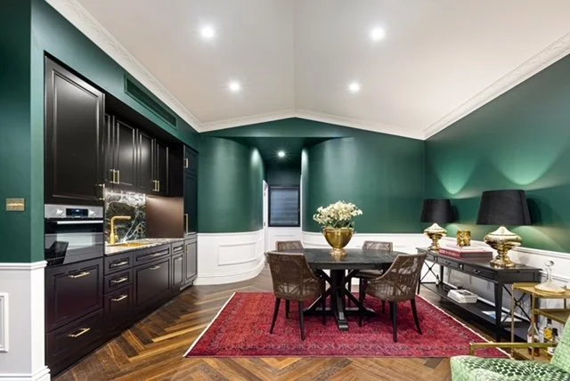 A dining room with dark green walls, black cabinetry, and a marble kitchen backsplash. There is a round dining table with four chairs on a red area rug, and various decorative items including a vase of white flowers and gold accents.