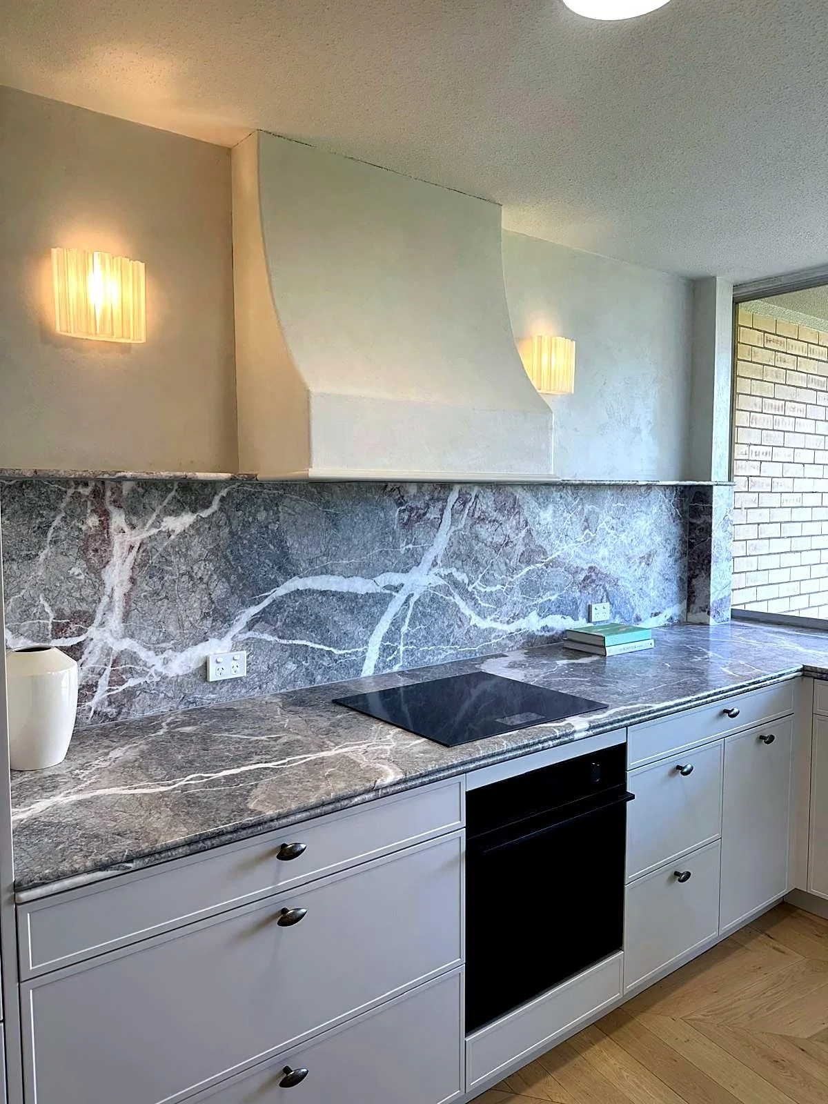 Kitchen with white cabinets, dark marble countertop, marble backsplash, electric cooktop, and decorative wall lamps.