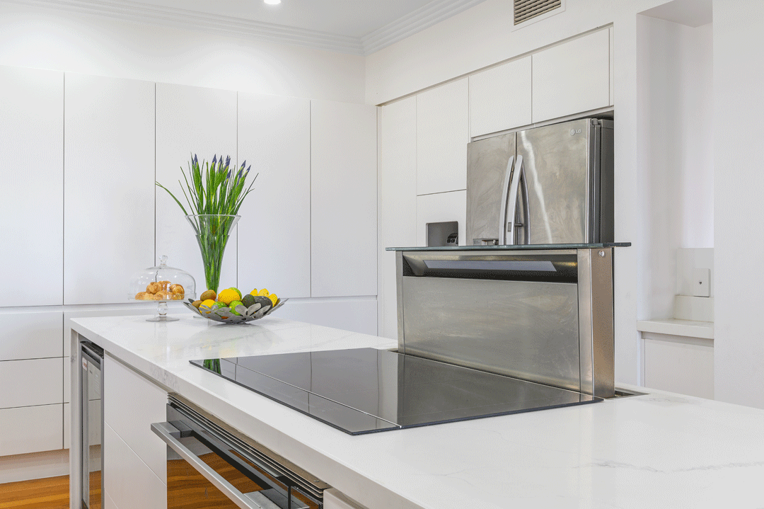 Modern kitchen with white cabinets, a white countertop, a stove, a stainless steel refrigerator, a vase with green and purple flowers, a bowl of lemons, and a glass jar of cookies.