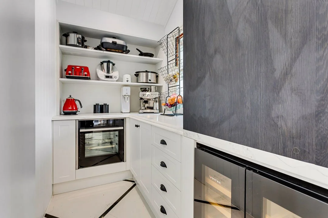 Small kitchen with white cabinets, open shelves with kitchen appliances, black handles on drawers, a window, and a black wall clock.