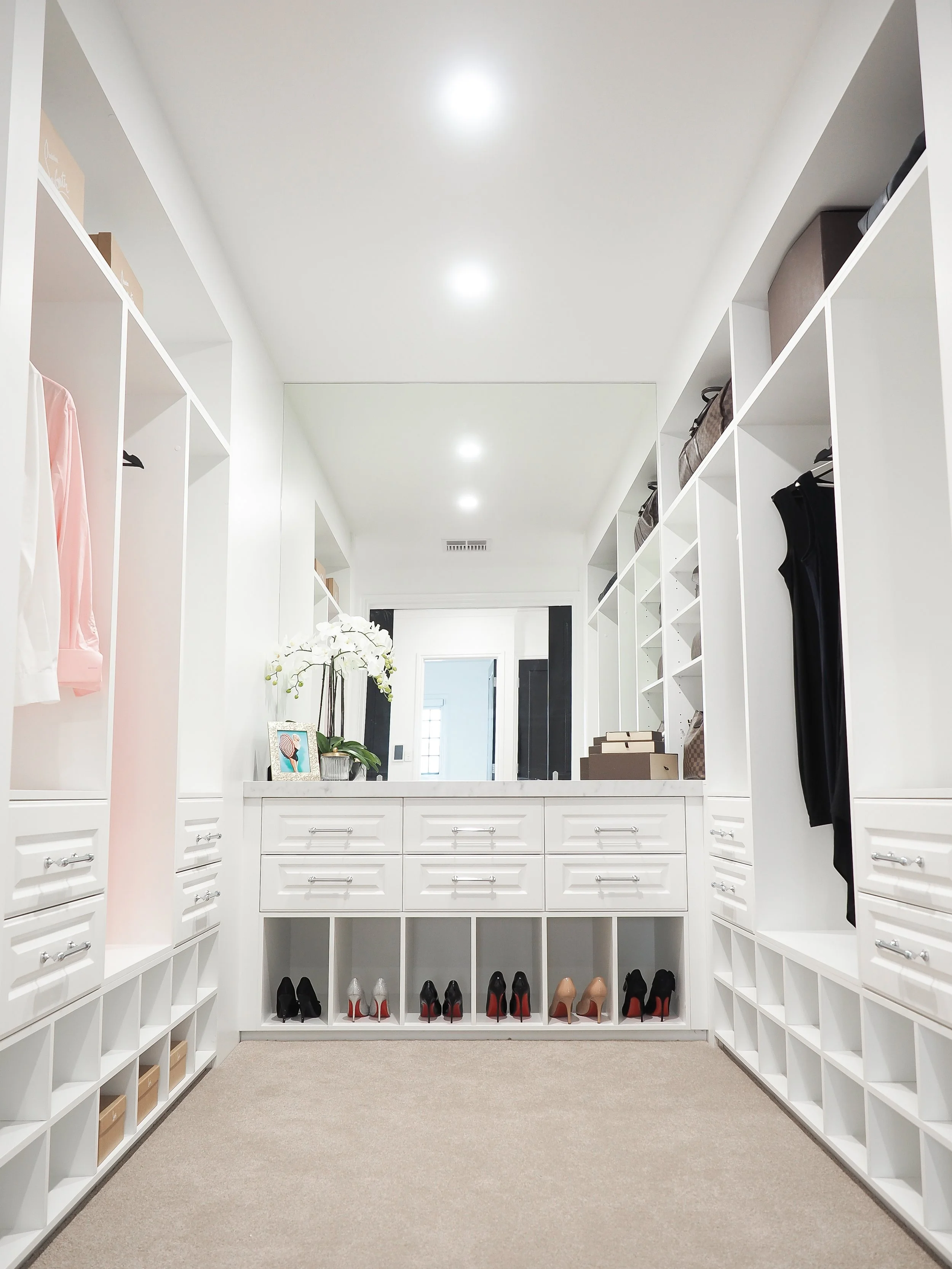 A spacious walk-in closet with white shelving units on both sides, containing shoes, handbags, and clothing. A pair of shoes and accessories are arranged at the bottom, with decorative items and a picture on the left side. Recessed ceiling lights ill