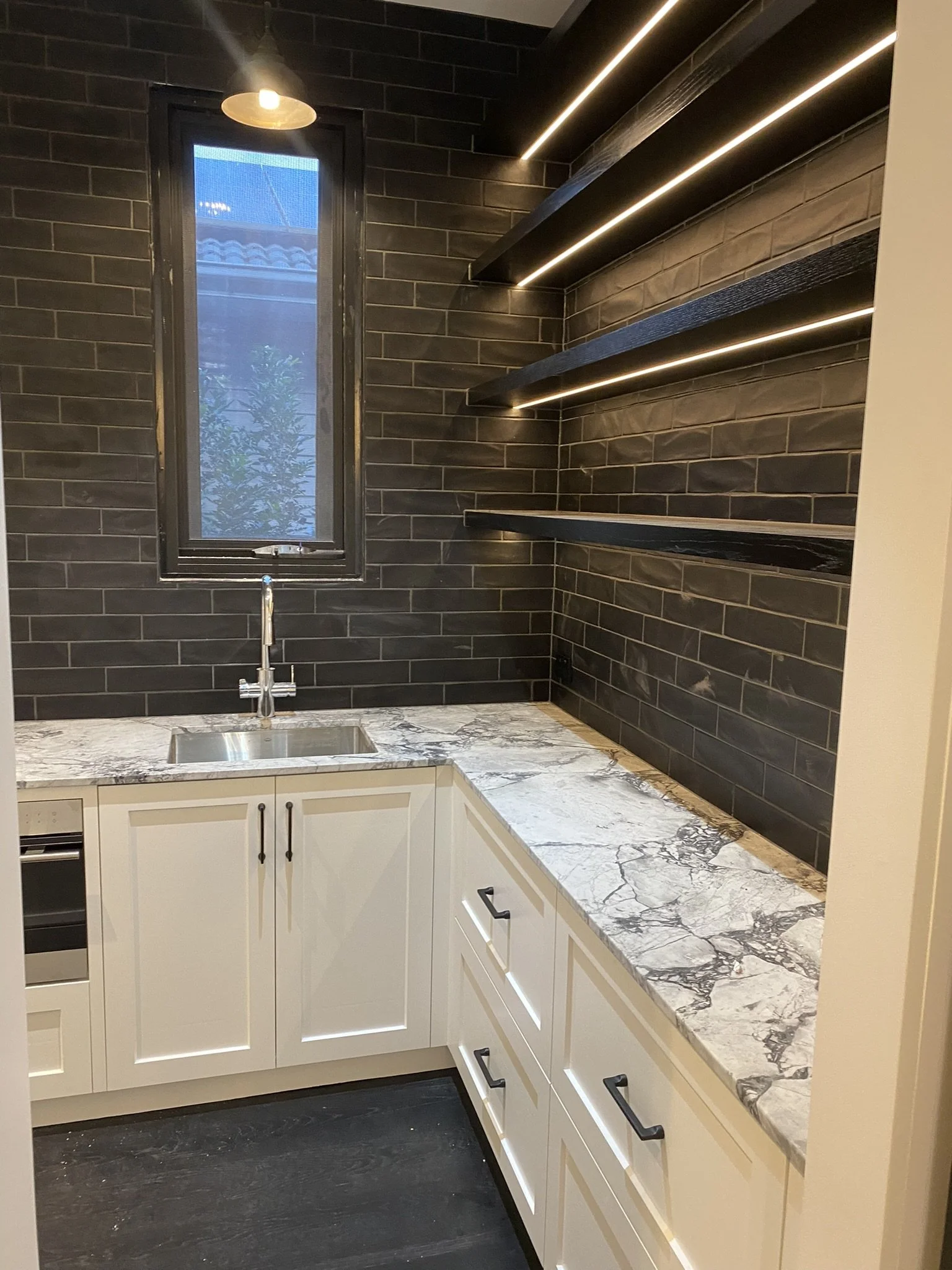 A modern kitchen with white cabinets, a marble countertop, a small sink, black hardware, dark wood flooring, a black tiled wall, a window, and black shelves with lighting.