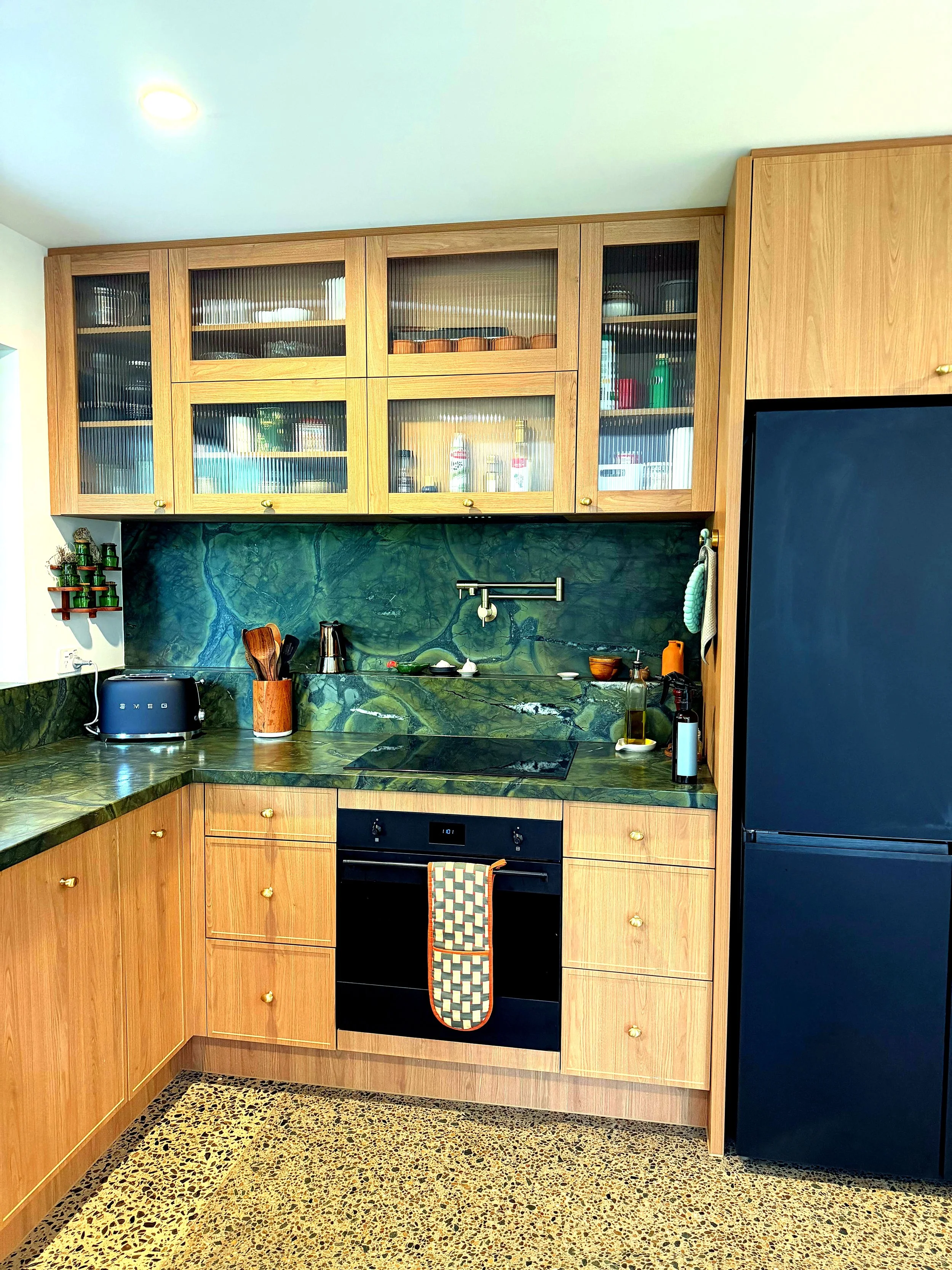 Kitchen with wooden cabinets, a green marble backsplash, a black oven with a checkered oven mitt hanging, a black refrigerator, and small plants on the countertop.