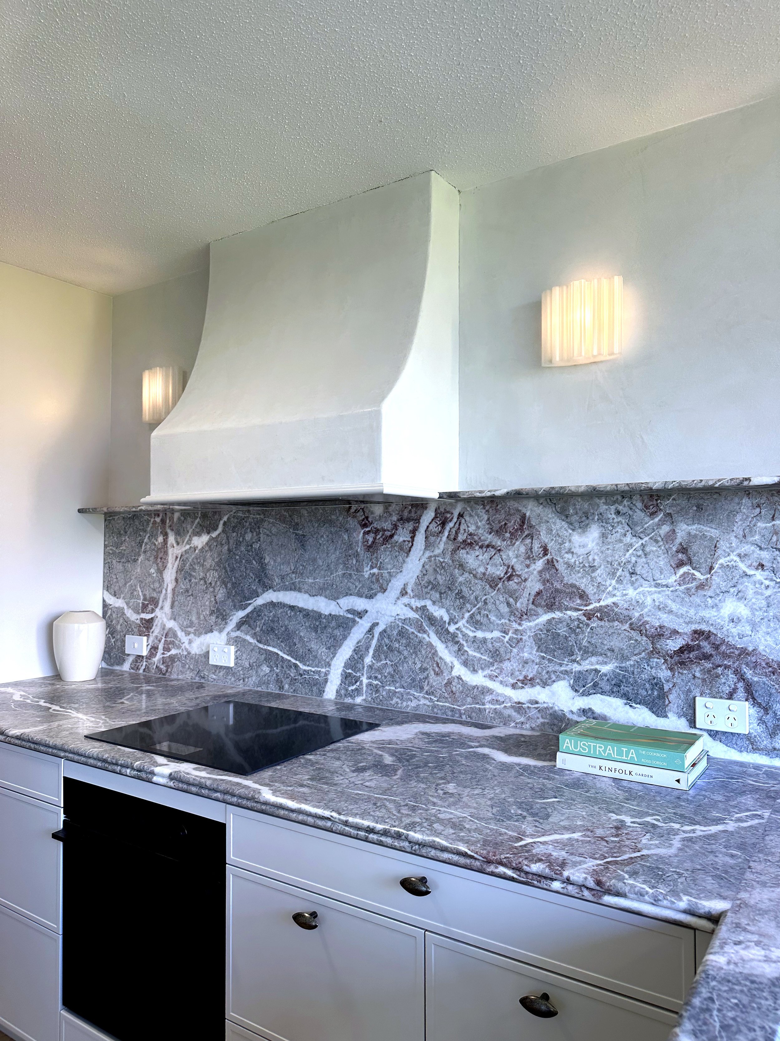 A kitchen with a marble countertop, a black stove, and a white cabinet. There are three books titled 'Australia,' 'The Cook Book,' and 'The Kinfolk Garden' on the counter. Two wall-mounted light fixtures are visible, and a tall white vase is on the l