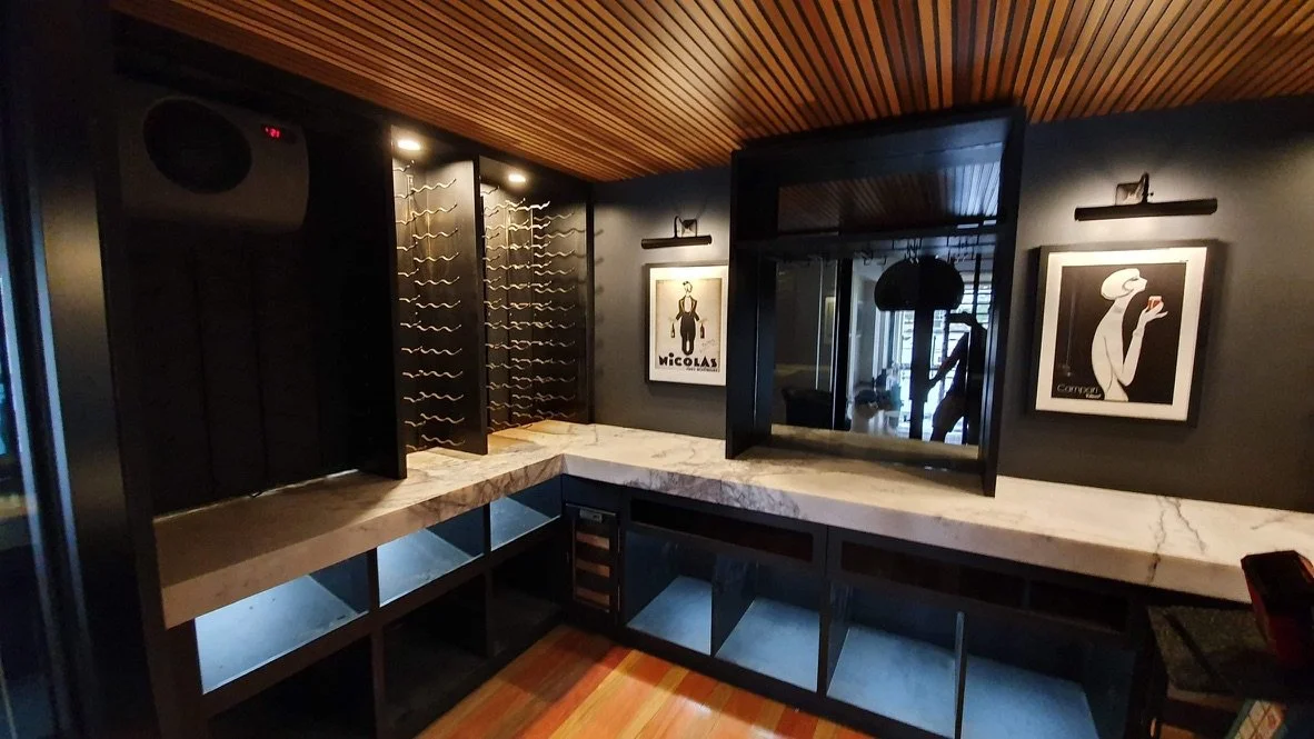 A wine storage area with a marble countertop, wine racks, framed art on dark wall, mirror, and wood ceiling.
