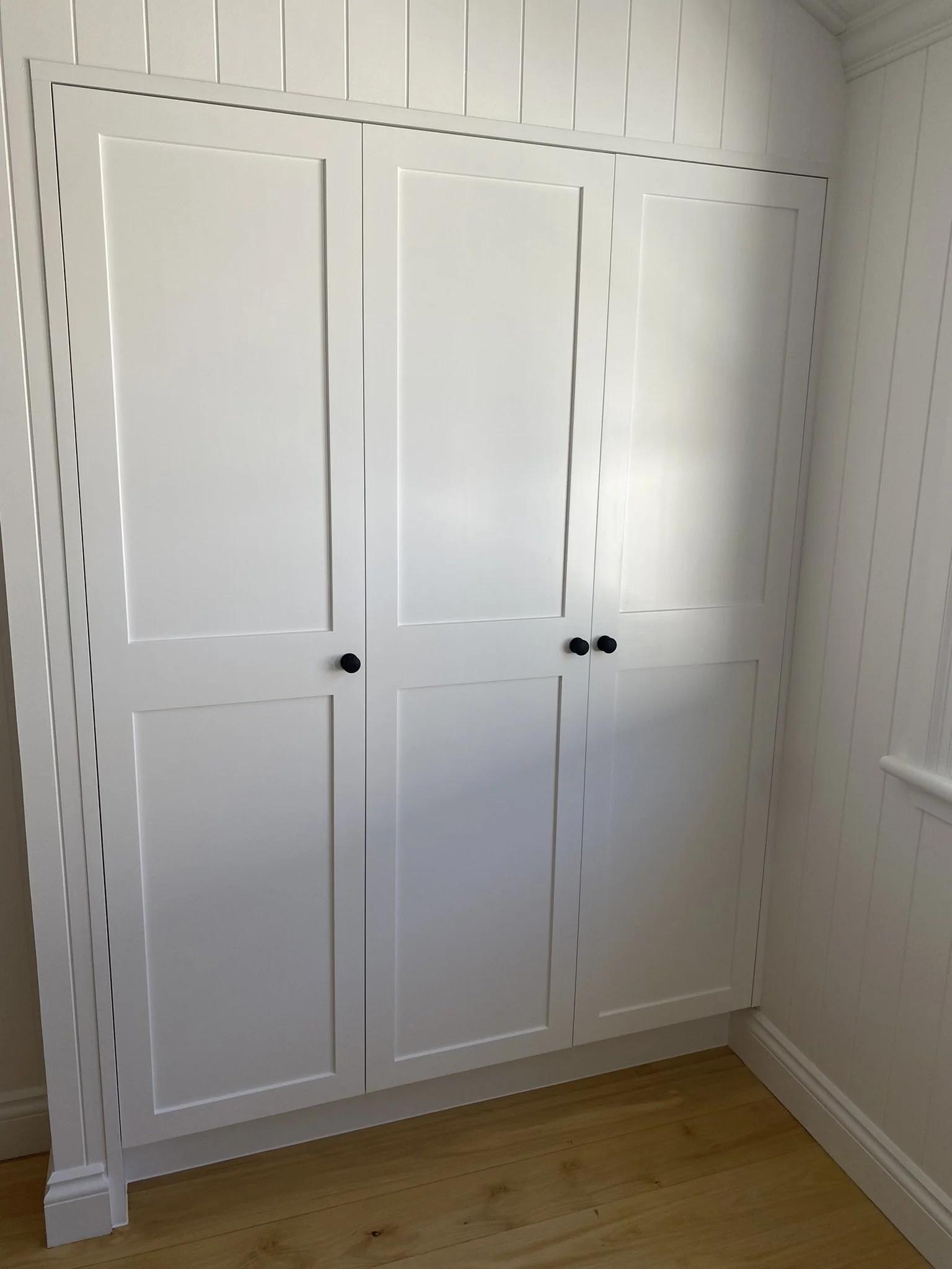 White kitchen cabinet with black knob handles, wooden floor, and beadboard wall