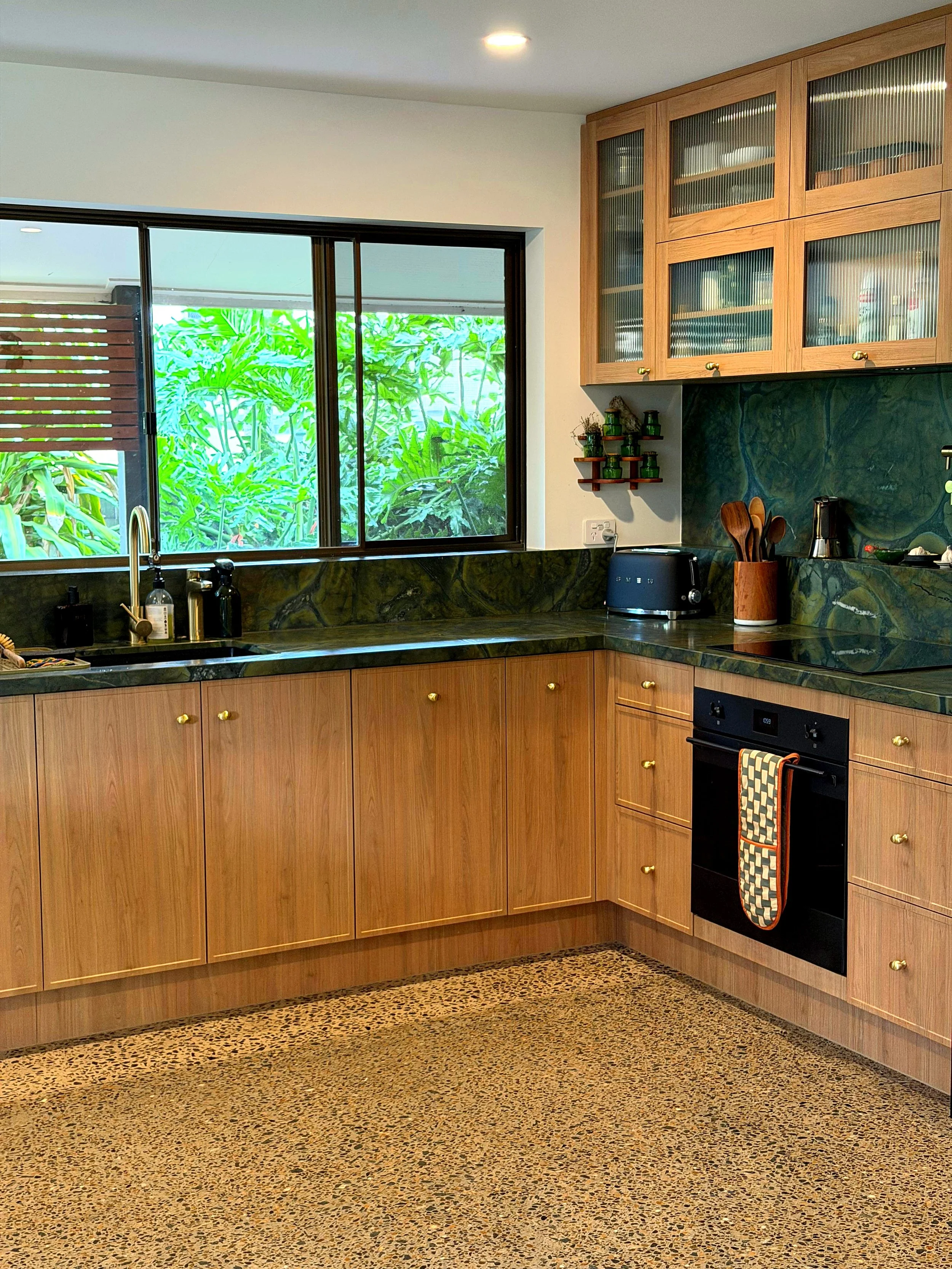 Kitchen with wooden cabinets, green marble countertops and backsplash, window overlooking greenery, black oven with an orange and brown oven mitt, black toaster, and kitchen utensils in a wooden container.