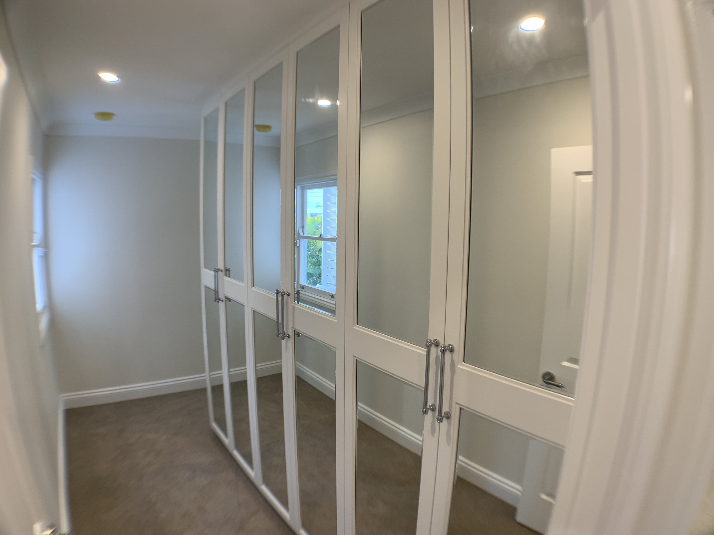 A closet with white cabinets and mirrored doors, a window with greenery outside, and ceiling lights.