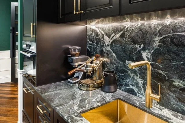 Close-up of a modern kitchen countertop with a black marble backsplash, gold sink faucet, and coffee brewing equipment on the dark cabinet with gold handles.
