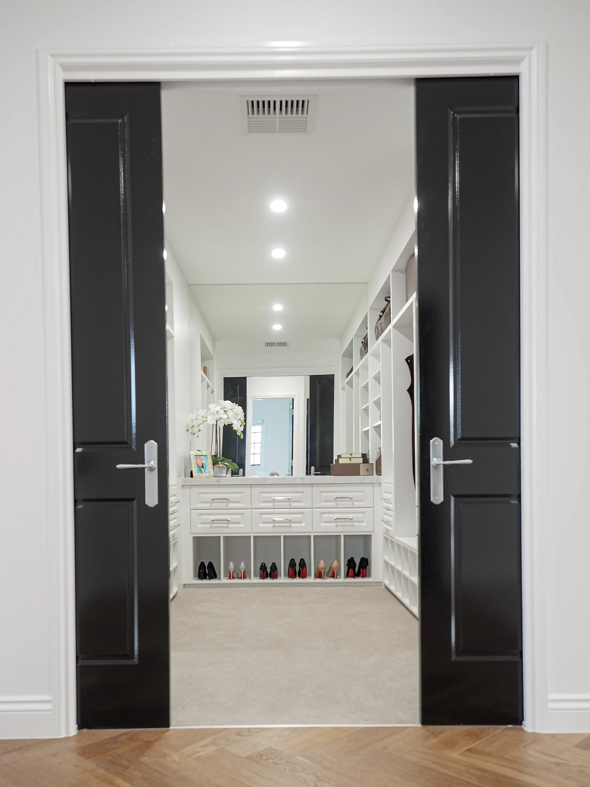 View of a walk-in closet seen through an open doorway with black doors, containing shelves with high heels at the bottom, a mirror, and decorative items.