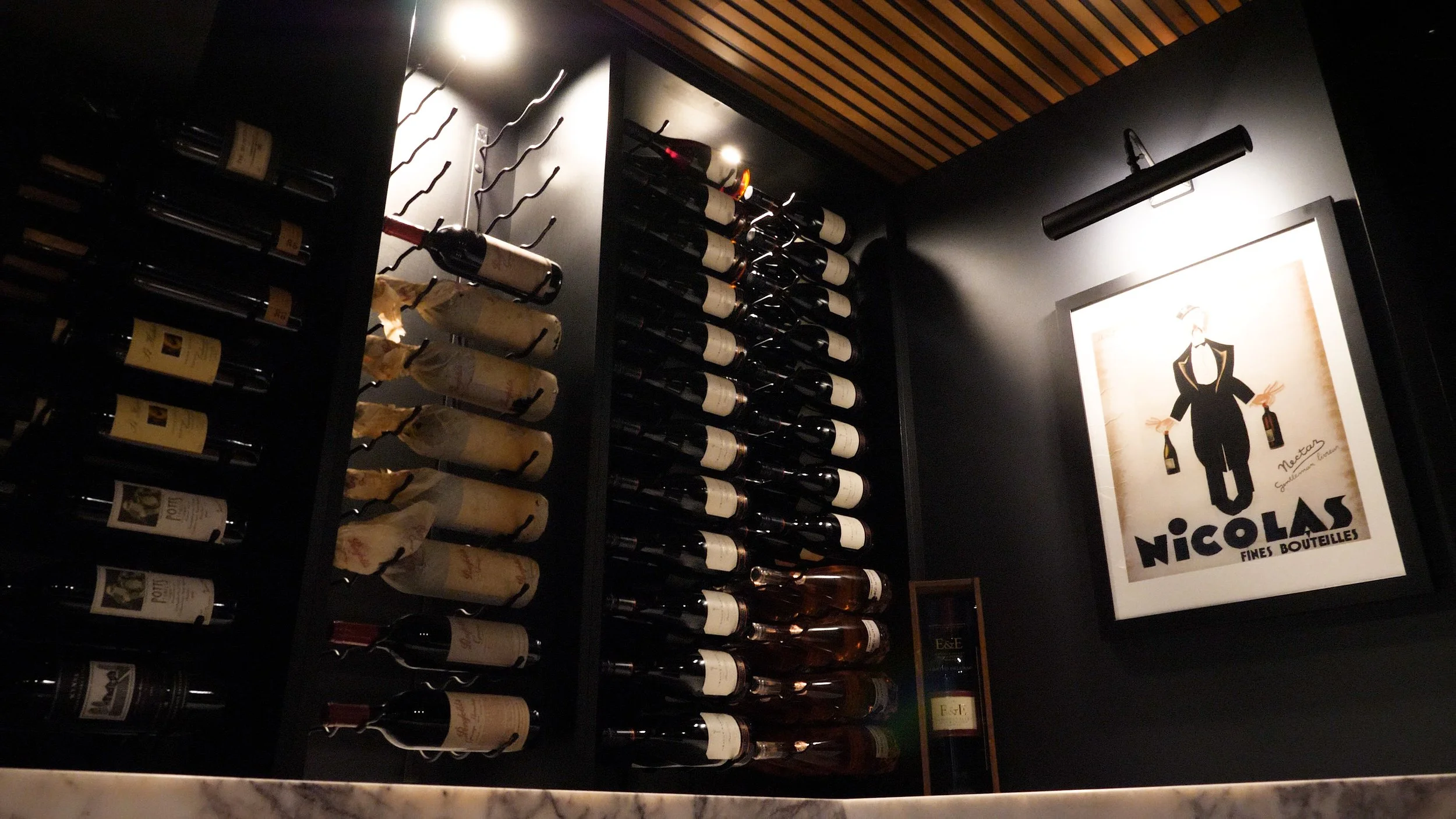 A wine cellar with shelves holding bottles of wine, some wrapped in paper. There is a framed poster of a cartoon sommelier holding wine bottles next to a spotlight.