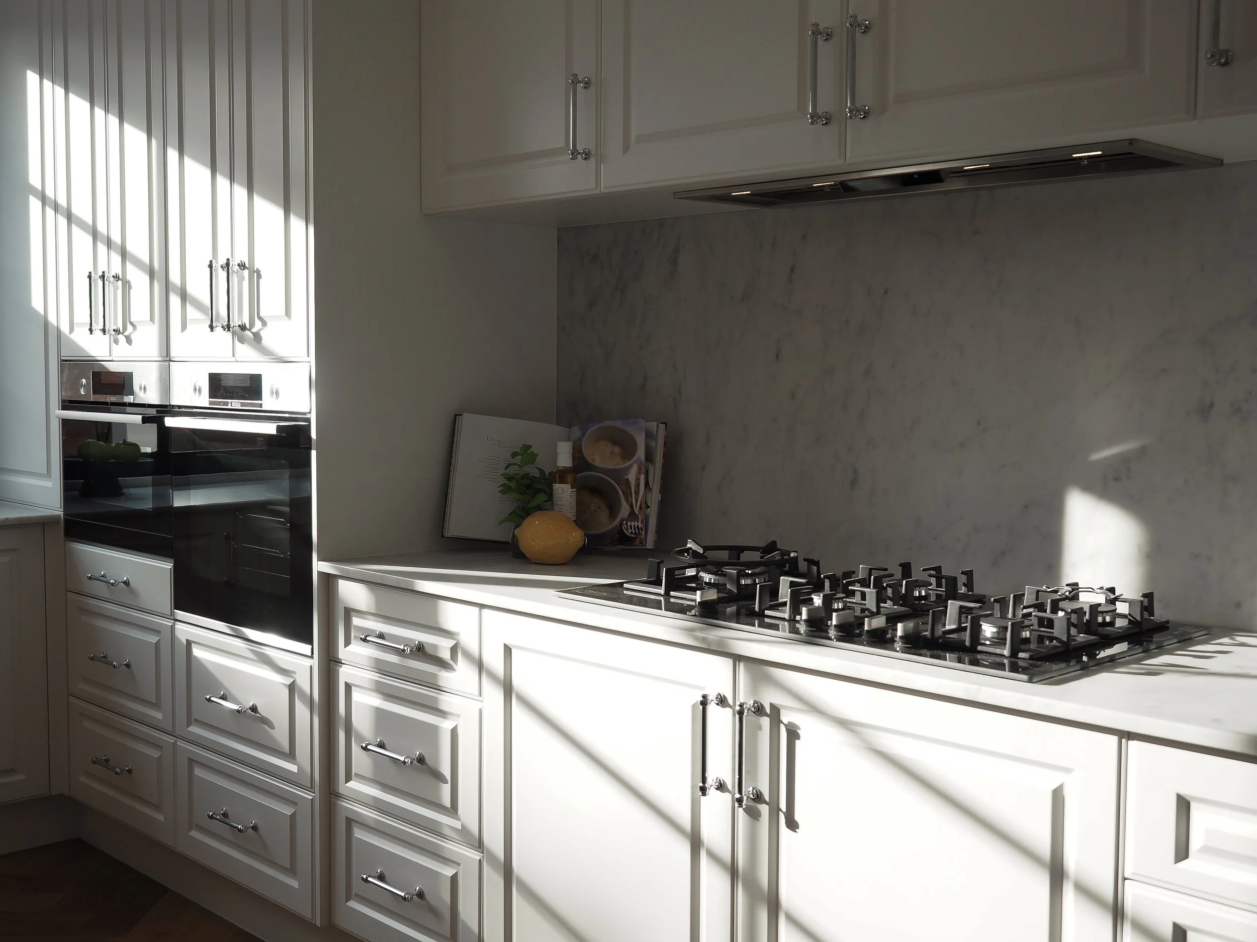 White kitchen cabinets with digital ovens, a gas stove, a marble backsplash, and countertop displays with a cookbook, plant, and condiments, with sunlight streaming through a window.