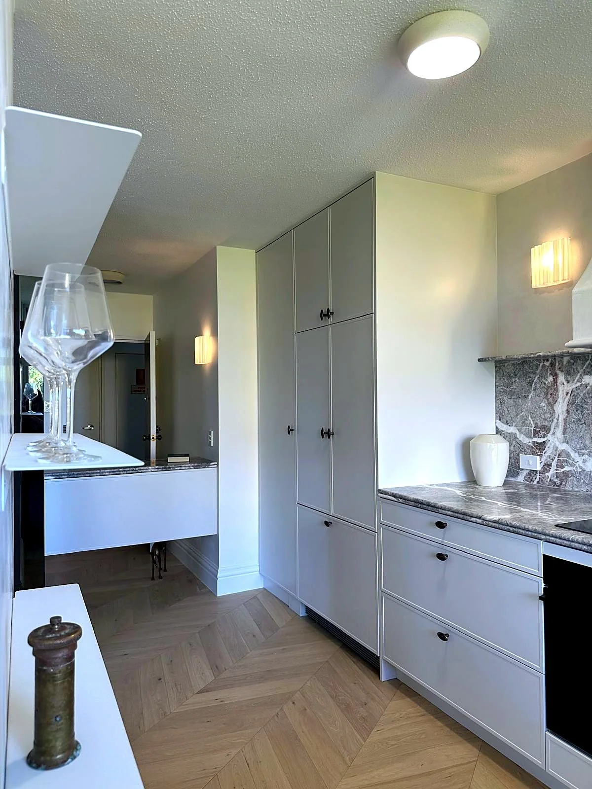 Modern kitchen with white cabinets, marble countertops, and a wood floor with a herringbone pattern. There are two glasses on a white shelf in the foreground, and a decorative vase on the counter near the wall. The ceiling has a textured finish and t
