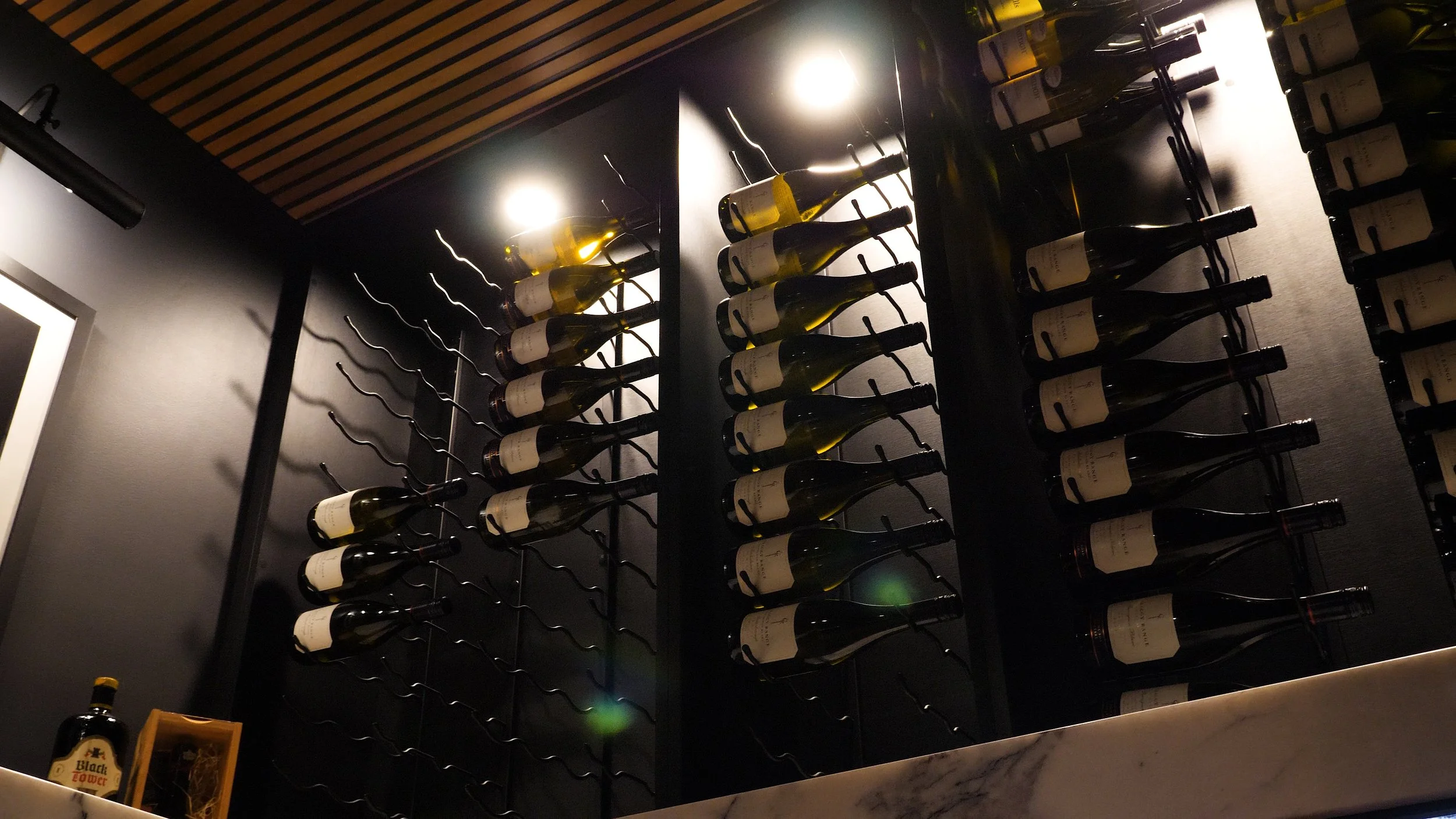 This custom wine room designed and built by Utopia Kitchens + Living brings a touch of the unexpected to a Brisbane home. Floor-to-ceiling black powder-coated metal wine rack systems are mounted against deep charcoal cabinetry panels, creating a dram