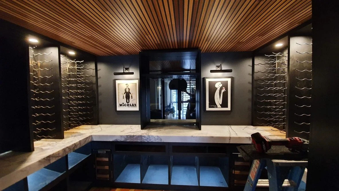 Wine storage area with wine racks on the walls, framed artwork between the racks, and a marble countertop on the bottom with shelves underneath. There is a door in the center leading to another room, a wooden ceiling, and some lighting fixtures.