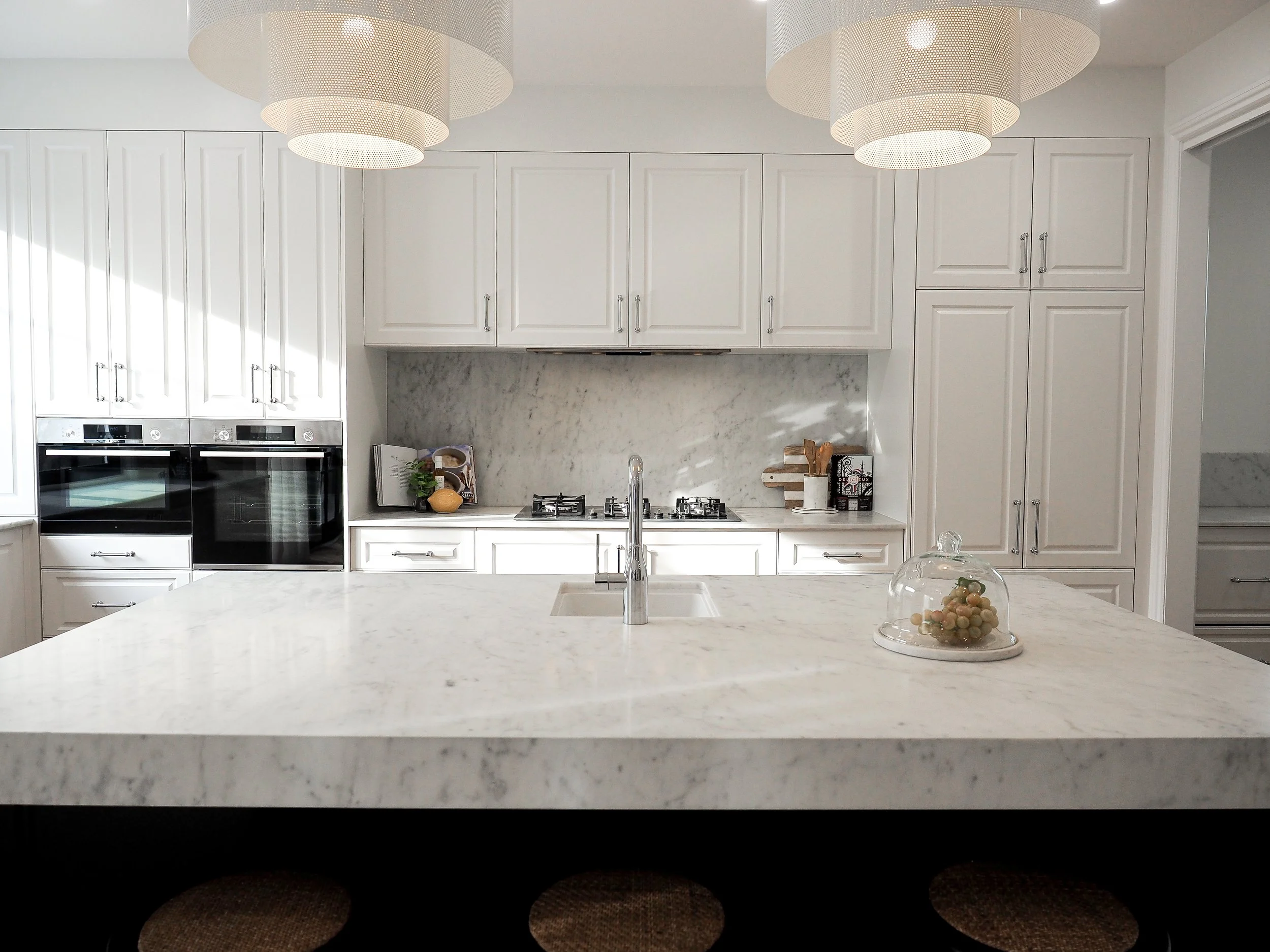 This Hampton style kitchen renovation in Ascot, Brisbane showcases the finest details of classic Australian luxury living. The cabinetry features full-height profiled shaker doors in a warm white finish, complemented by polished chrome bar handles th