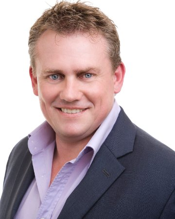 Professional headshot of a man with light brown hair, blue eyes, wearing a dark suit jacket and a light purple dress shirt, smiling against a plain white background.