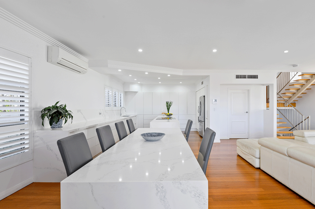 Modern white kitchen and living area with a marble dining table, gray chairs, white sofa, staircase, and wooden floors.