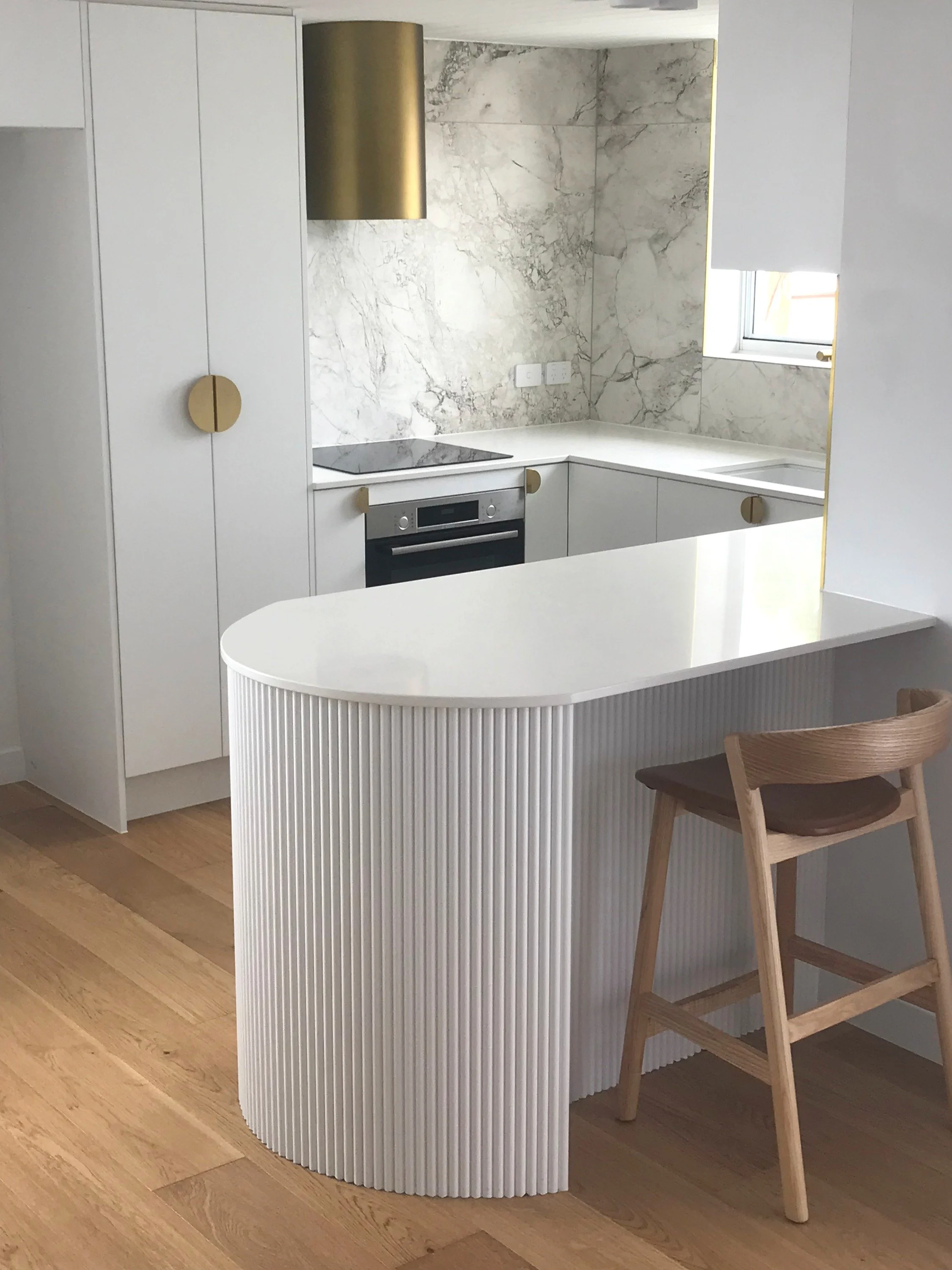 Modern kitchen with white cabinetry, marble backsplash, built-in oven, induction cooktop, and a marble countertop. A small window is above the sink, and there's a wooden chair at a white, curved, fluted breakfast bar with a gold trim.