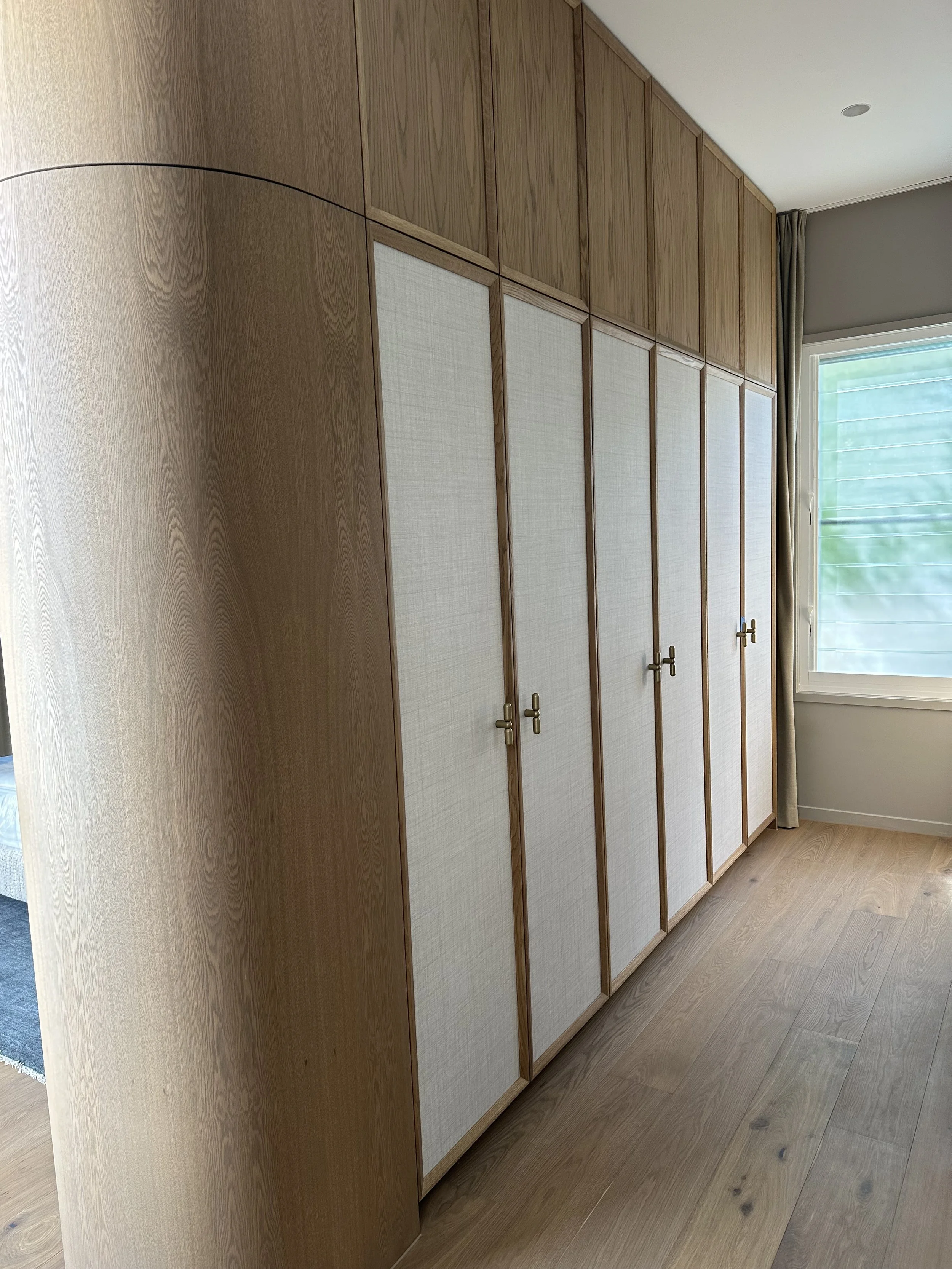 A row of wooden cabinets with light-colored doors and brass handles next to a window with curtains.