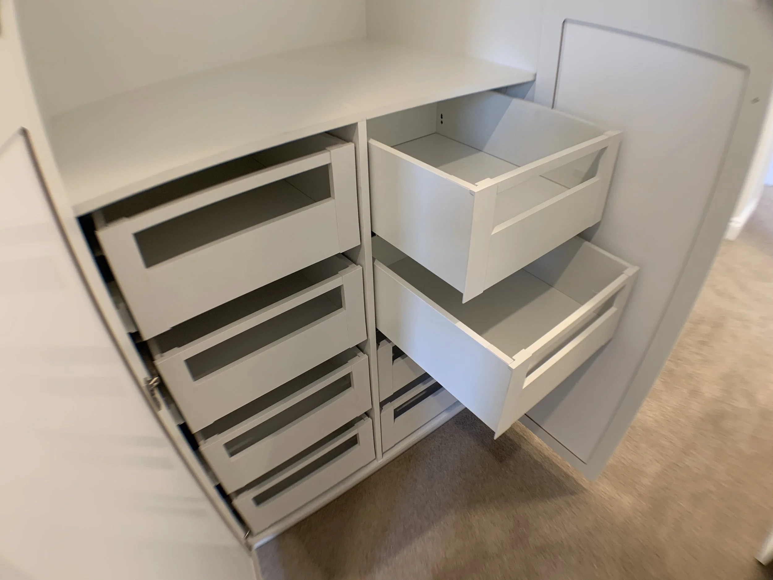 White built-in cabinet with multiple open drawers and shelves, some drawers are empty, located on a beige carpeted floor.