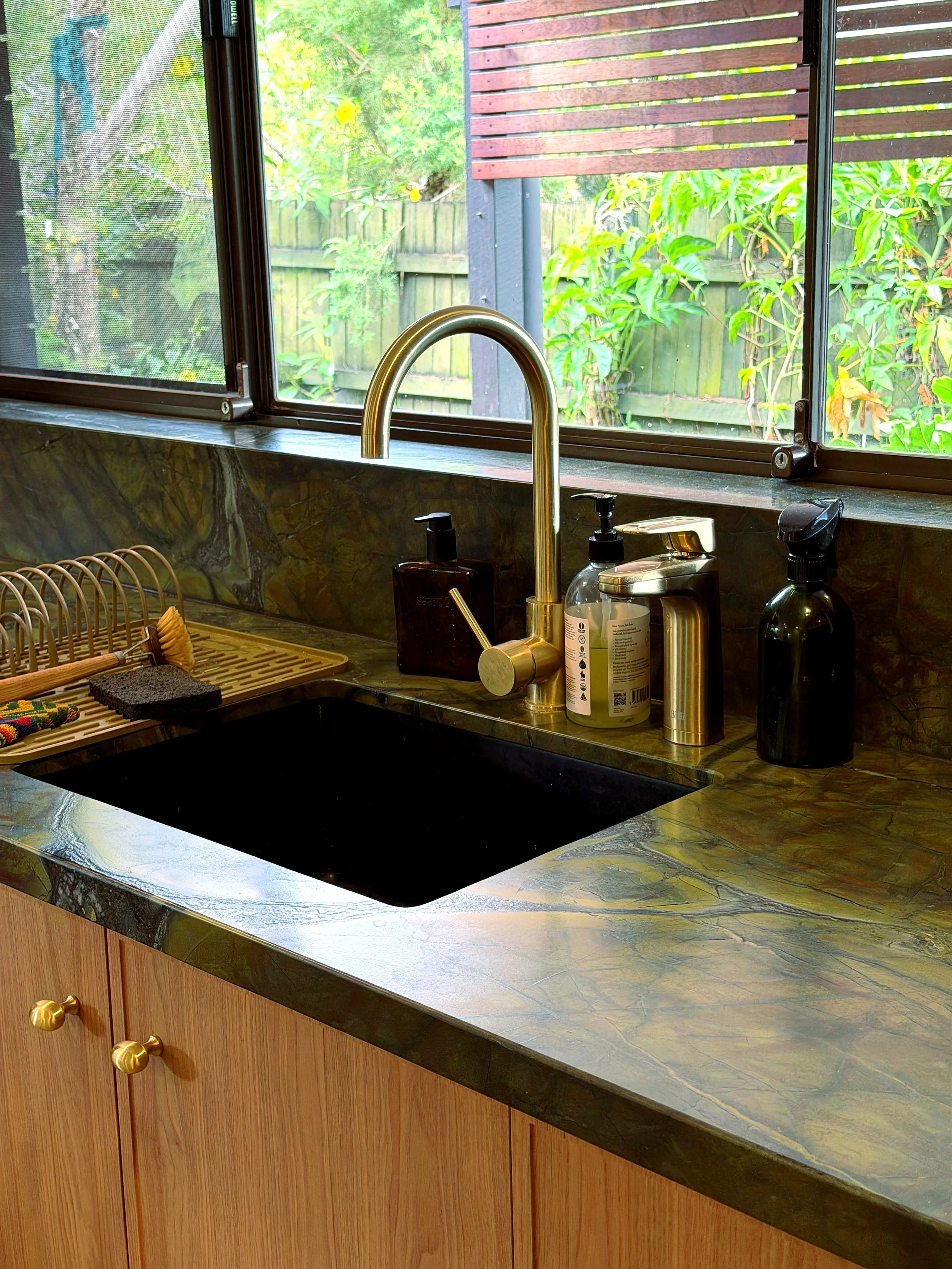 A kitchen countertop with a black sink, a gold faucet, three soap dispensers, a dish drying rack, a scrub brush, and a window with a view of a garden with trees and a wooden fence.